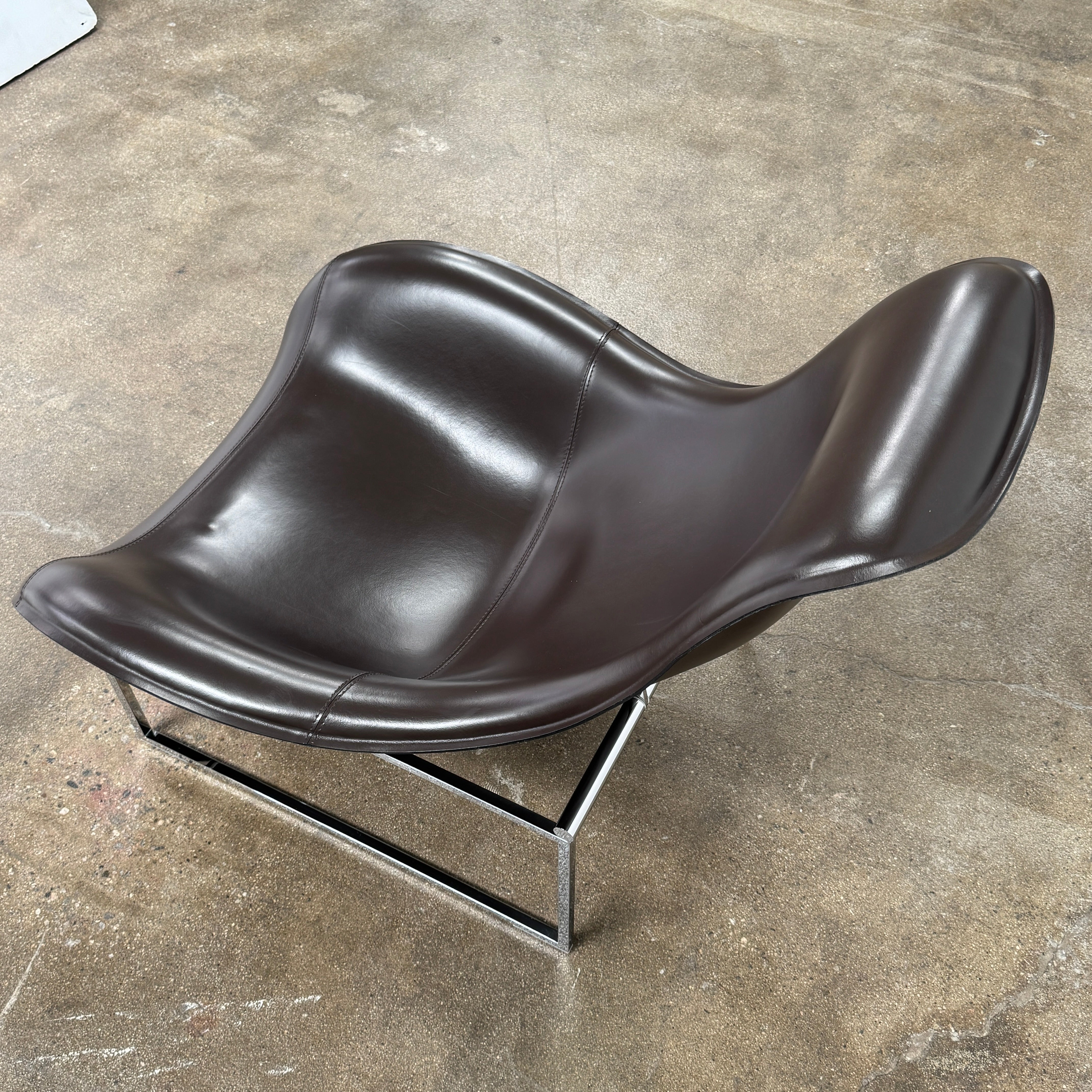 A B&B Italia Mart Relax Armchair with Tilting Mechanism in black saddle leather features a modern, curved design and is set on a polished concrete floor against a plain light gray wall.