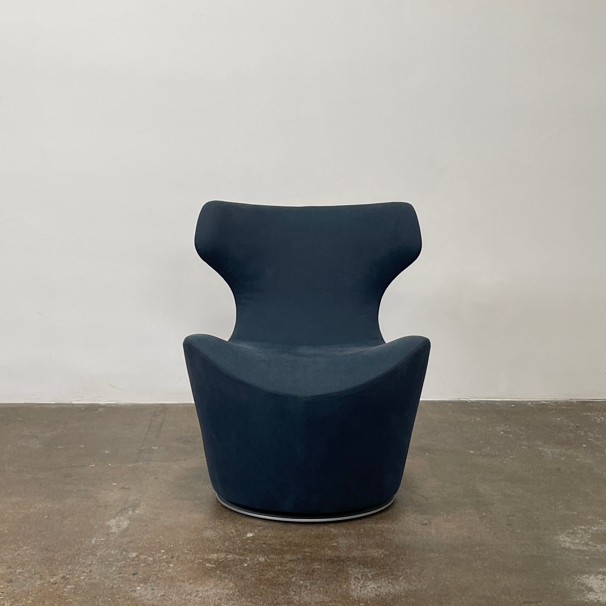 The B&B Italia Piccola Papilio Swivel Armchair is a modern piece with an abstract form and high curved backrest. Made from teal fabric, it stands on a swivel base against a plain white background.