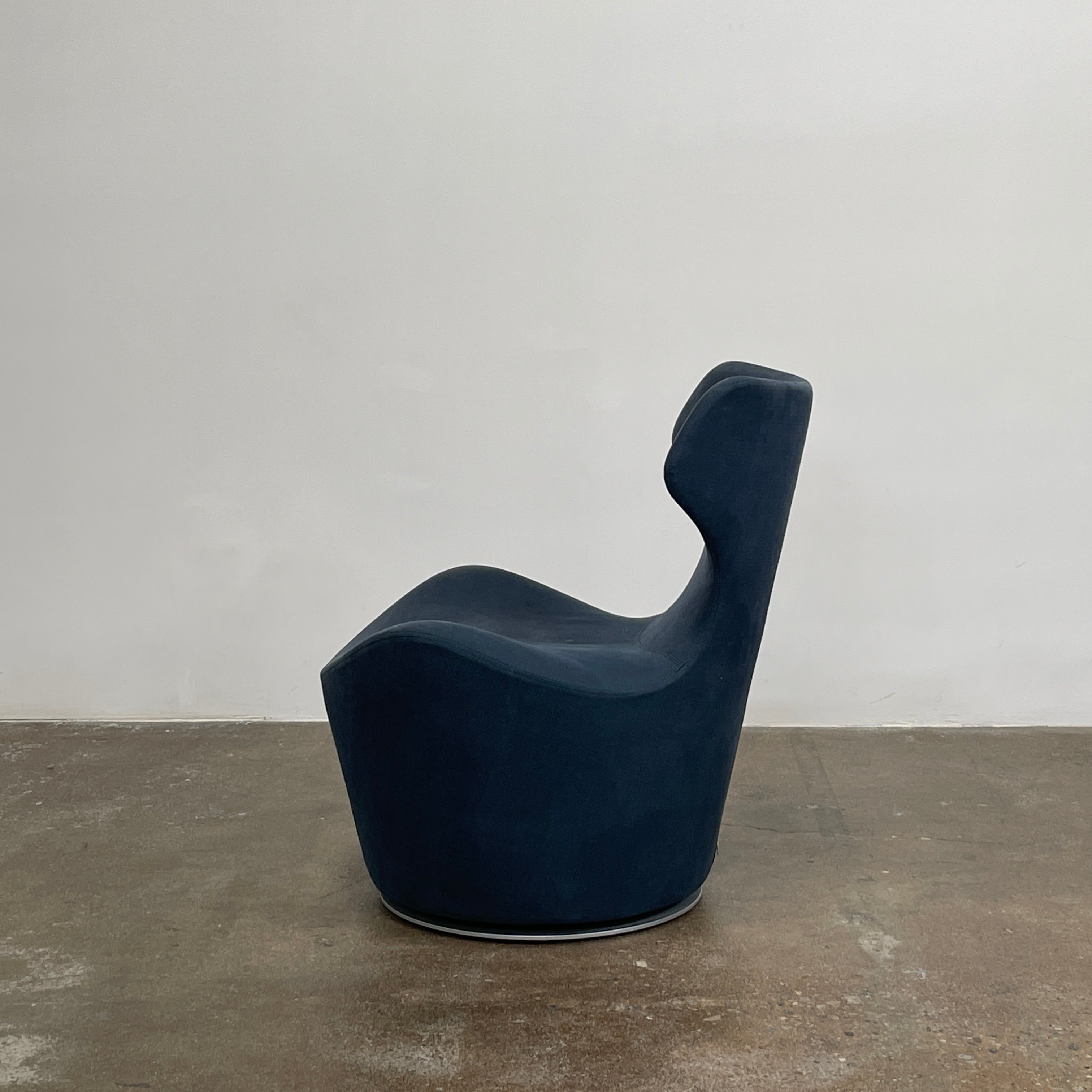 The B&B Italia Piccola Papilio Swivel Armchair is a modern piece with an abstract form and high curved backrest. Made from teal fabric, it stands on a swivel base against a plain white background.