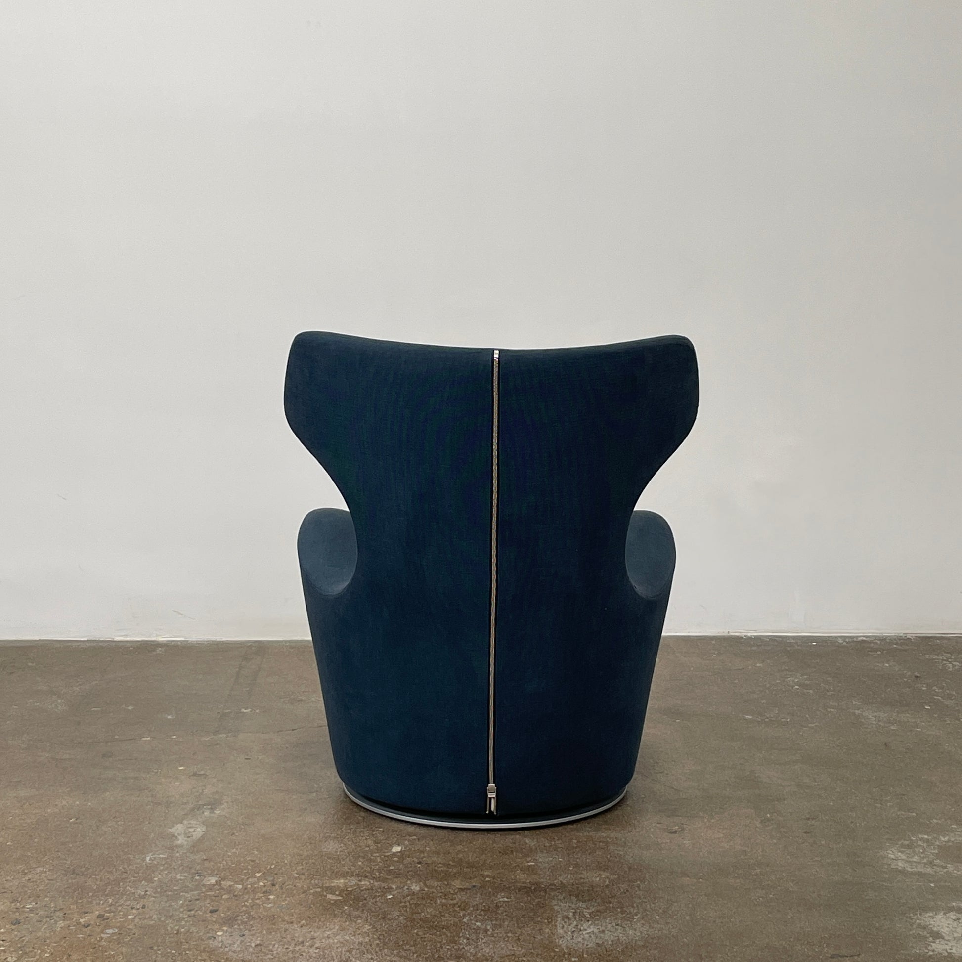 The B&B Italia Piccola Papilio Swivel Armchair is a modern piece with an abstract form and high curved backrest. Made from teal fabric, it stands on a swivel base against a plain white background.