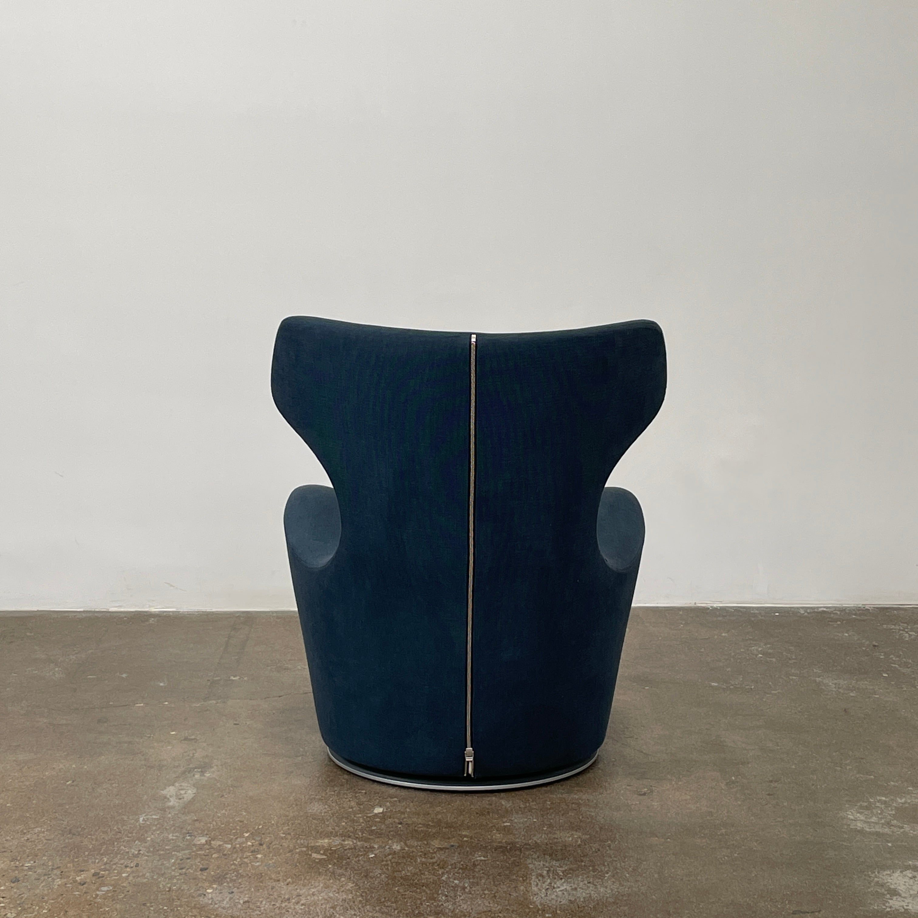 The B&B Italia Piccola Papilio Swivel Armchair is a modern piece with an abstract form and high curved backrest. Made from teal fabric, it stands on a swivel base against a plain white background.