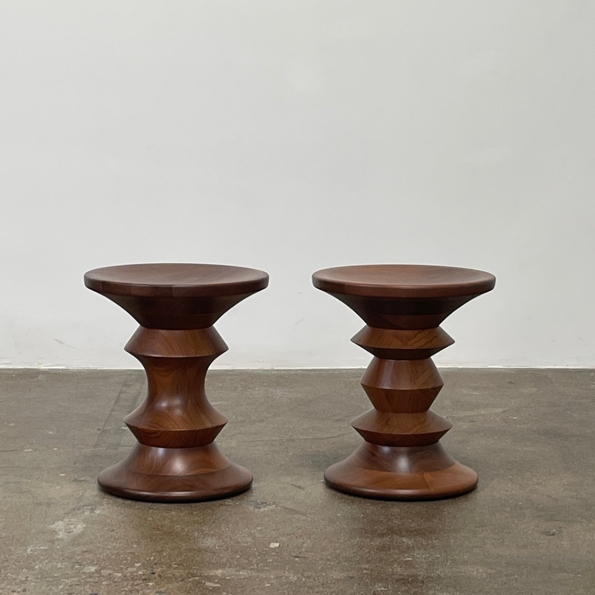The Eames Walnut Turned Shape C Stool by Herman Miller, featuring a solid-turned walnut design with a base resembling an abstract chess piece, stands on a concrete floor against a plain white wall.