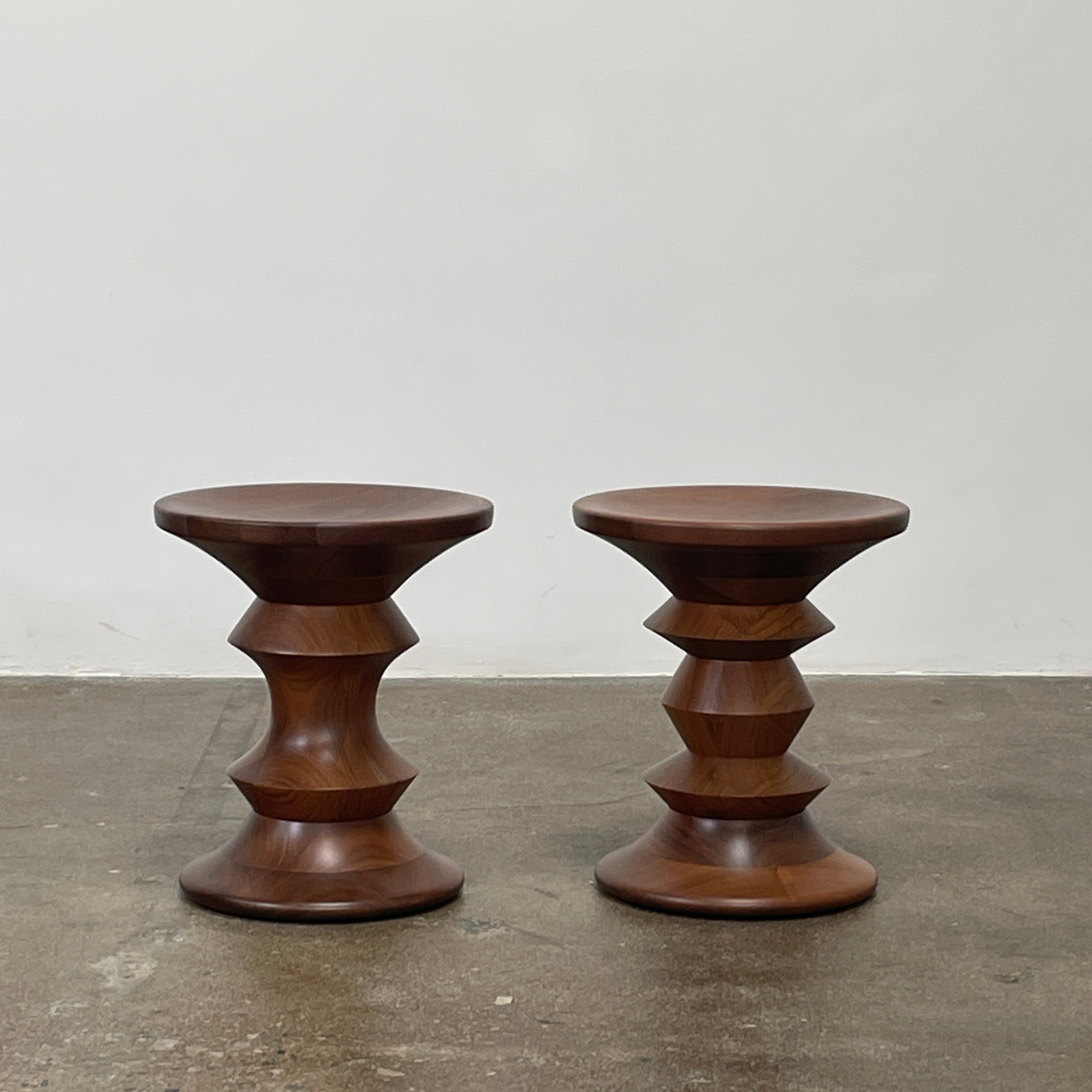 The Eames Walnut Turned Shape C Stool by Herman Miller, featuring a solid-turned walnut design with a base resembling an abstract chess piece, stands on a concrete floor against a plain white wall.