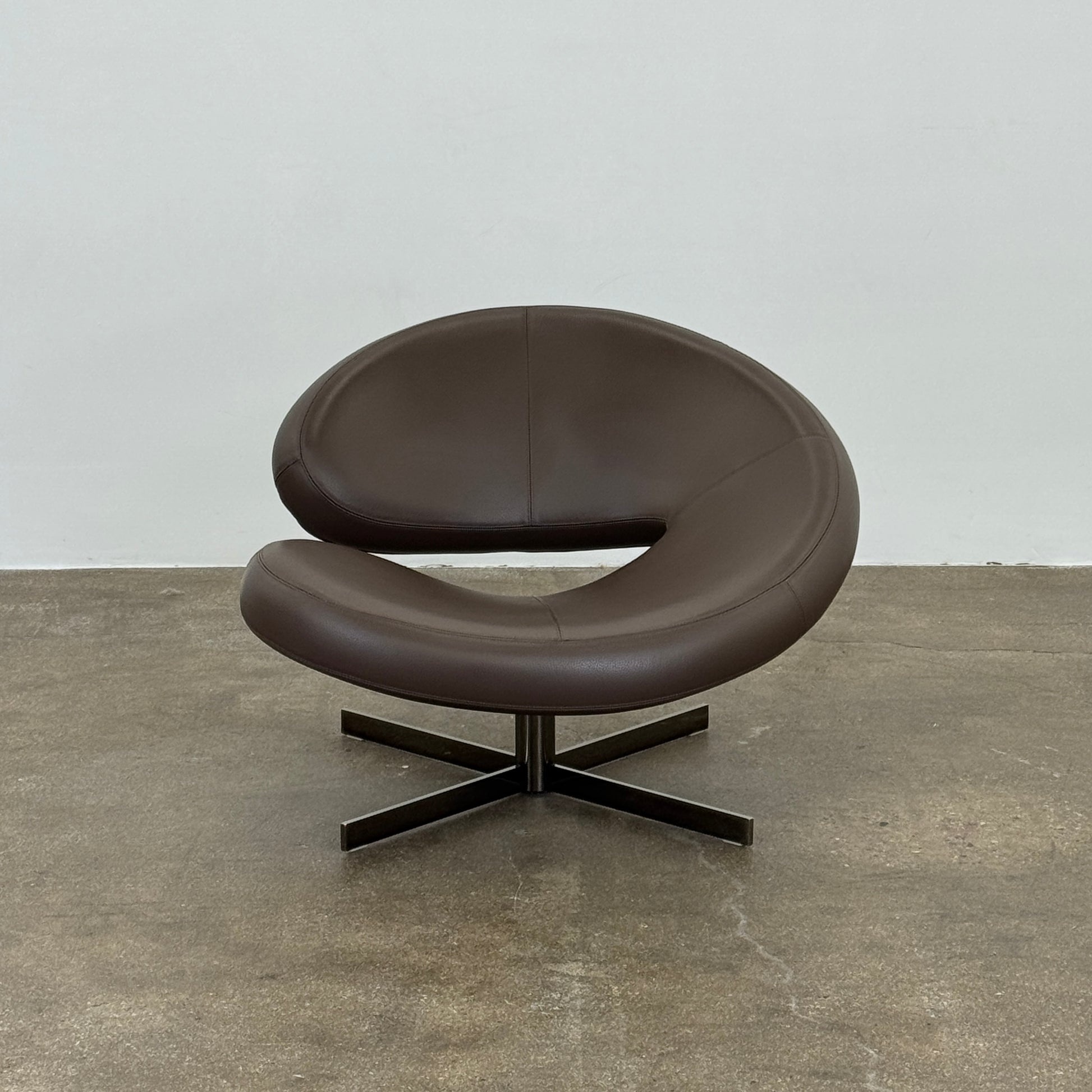 The Roche Bobois Nuage Swivel Chair is a modern, circular brown leather chair with a curved backrest and seat on a metal swivel base, set on a concrete floor against a plain white wall.