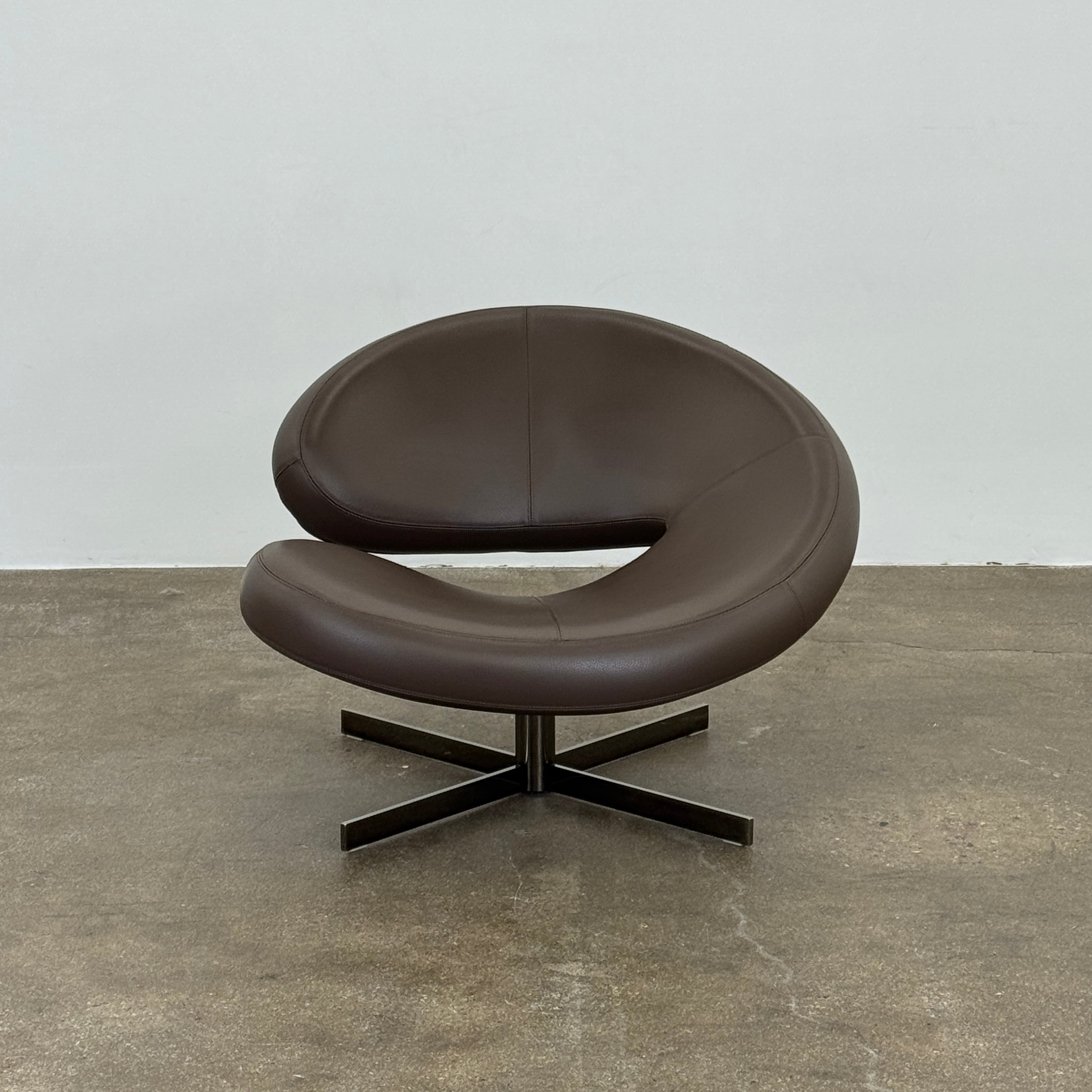 The Roche Bobois Nuage Swivel Chair is a modern, circular brown leather chair with a curved backrest and seat on a metal swivel base, set on a concrete floor against a plain white wall.