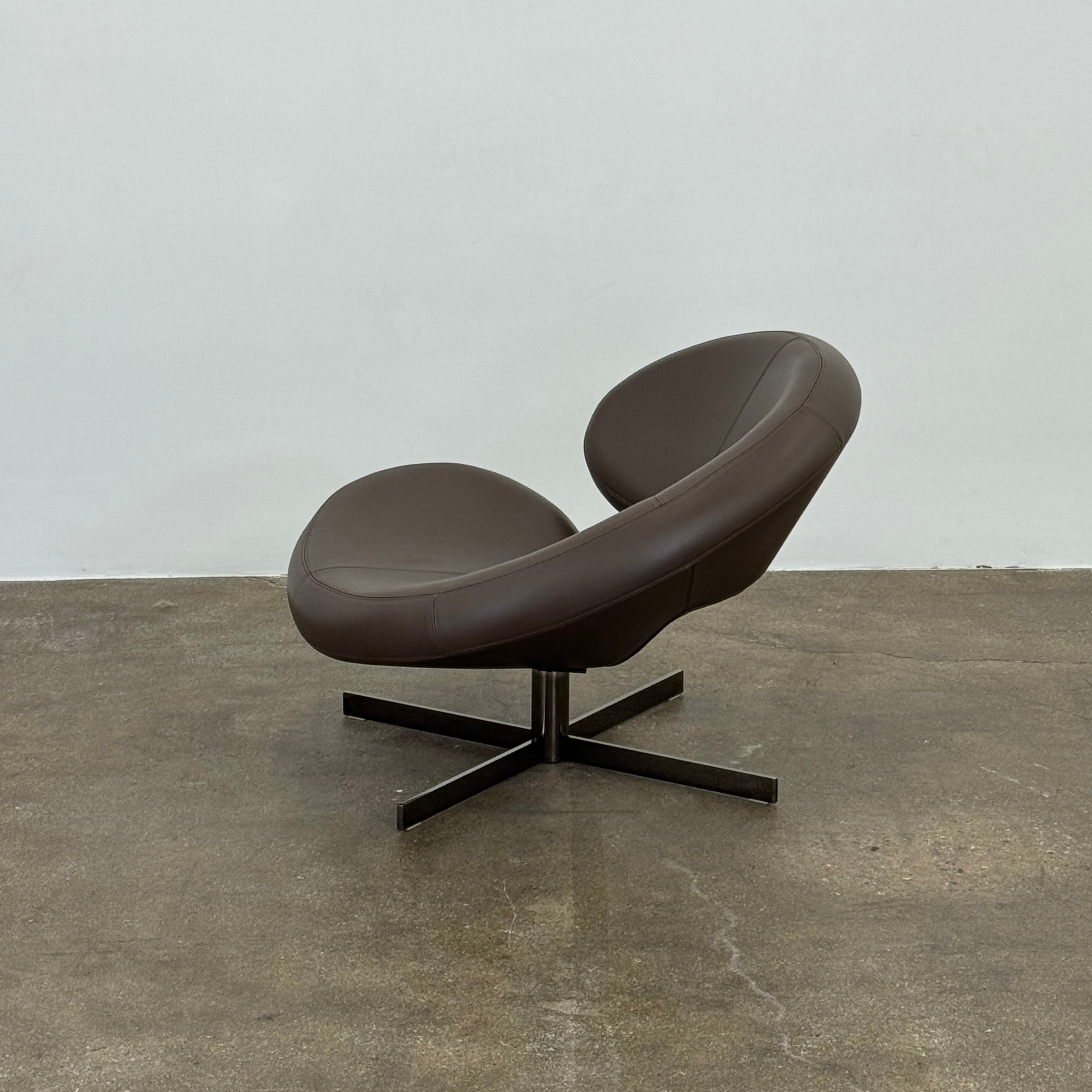 The Roche Bobois Nuage Swivel Chair is a modern, circular brown leather chair with a curved backrest and seat on a metal swivel base, set on a concrete floor against a plain white wall.