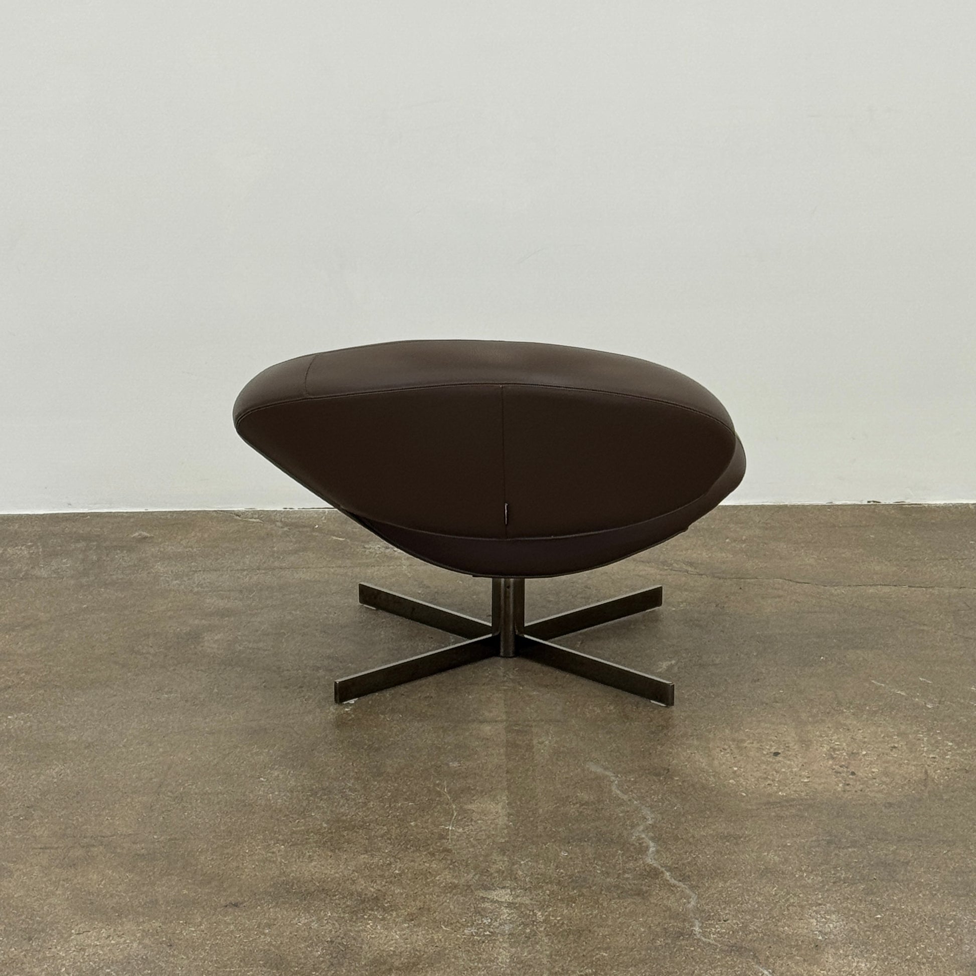 The Roche Bobois Nuage Swivel Chair is a modern, circular brown leather chair with a curved backrest and seat on a metal swivel base, set on a concrete floor against a plain white wall.