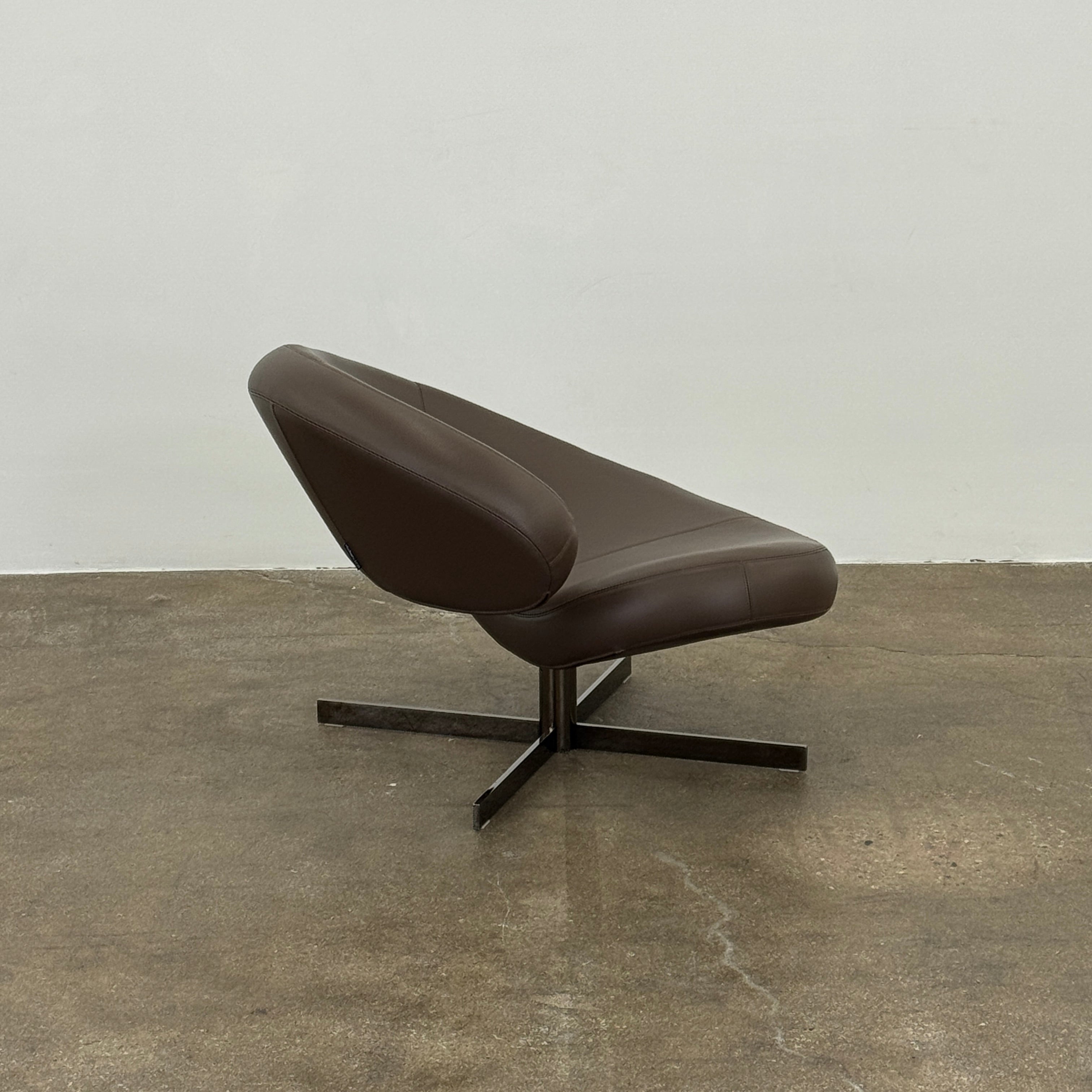 The Roche Bobois Nuage Swivel Chair is a modern, circular brown leather chair with a curved backrest and seat on a metal swivel base, set on a concrete floor against a plain white wall.