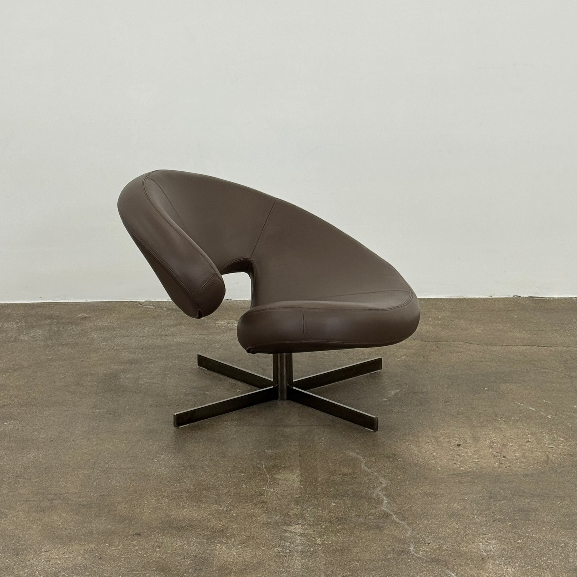 The Roche Bobois Nuage Swivel Chair is a modern, circular brown leather chair with a curved backrest and seat on a metal swivel base, set on a concrete floor against a plain white wall.
