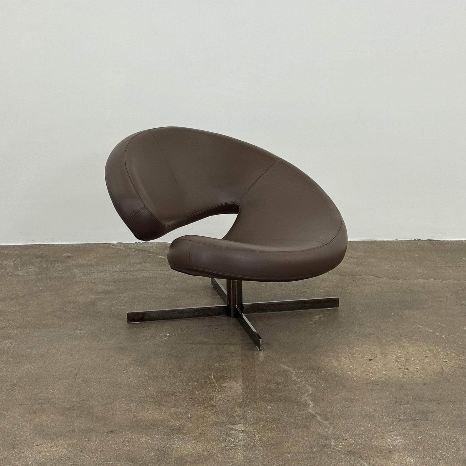 The Roche Bobois Nuage Swivel Chair is a modern, circular brown leather chair with a curved backrest and seat on a metal swivel base, set on a concrete floor against a plain white wall.