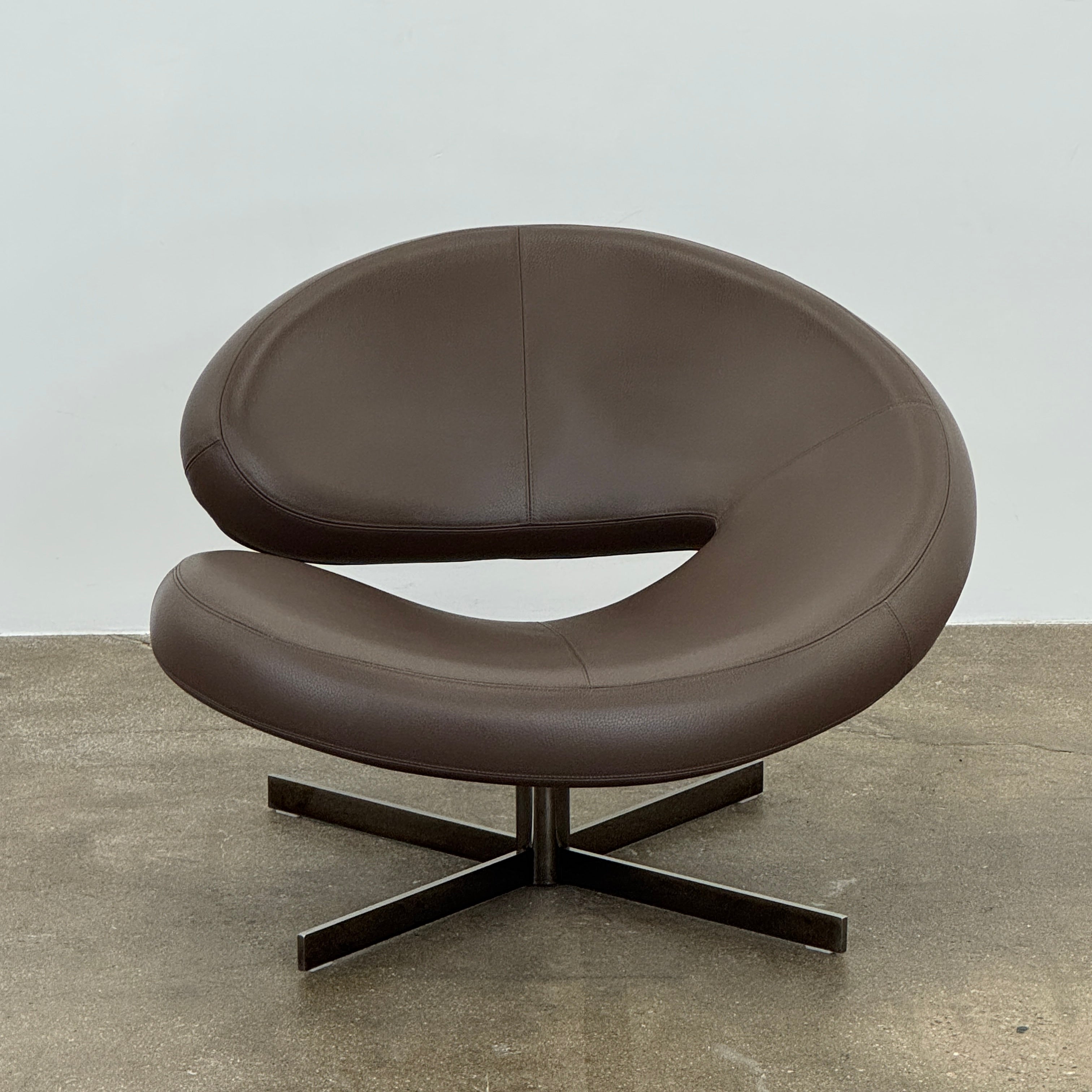 The Roche Bobois Nuage Swivel Chair is a modern, circular brown leather chair with a curved backrest and seat on a metal swivel base, set on a concrete floor against a plain white wall.