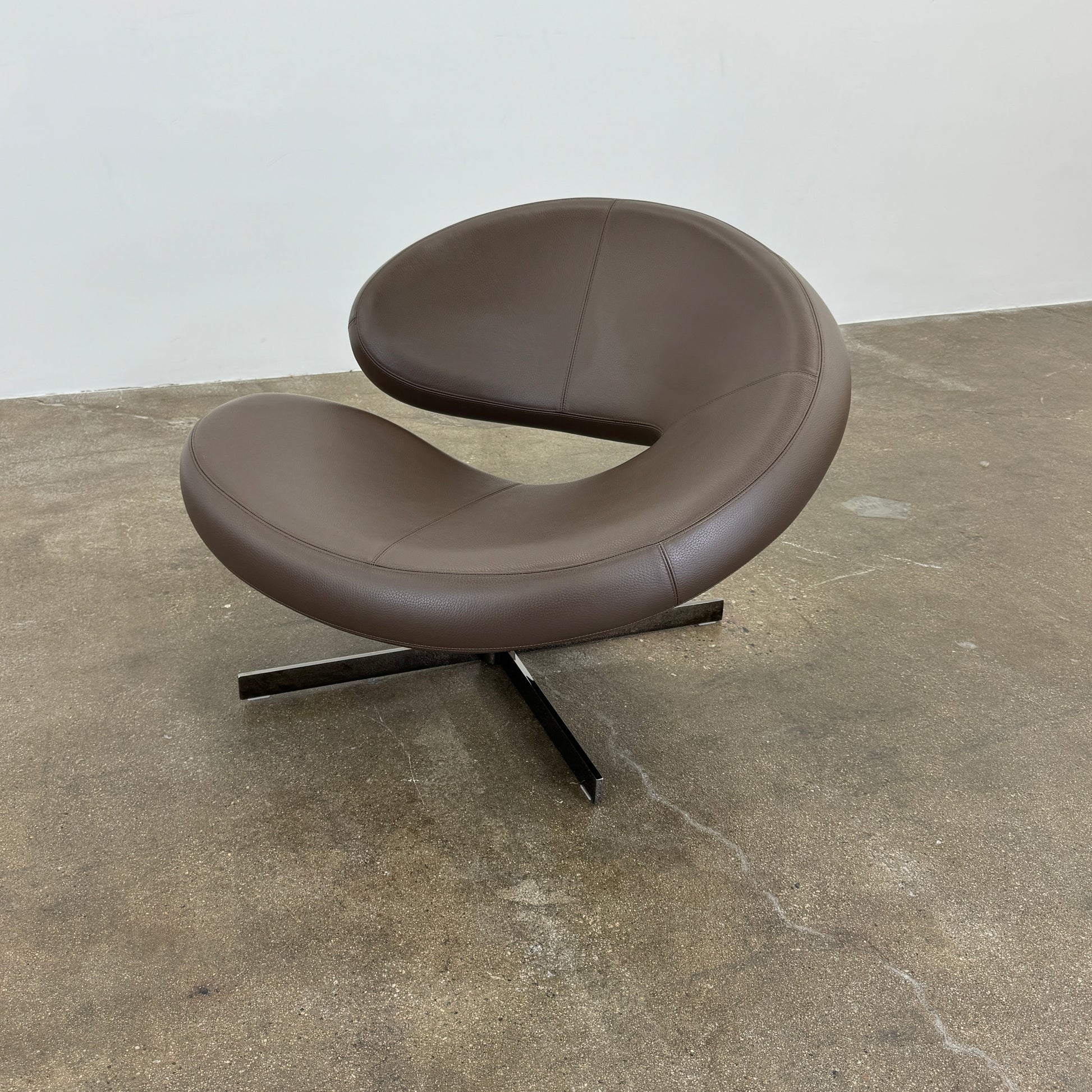 The Roche Bobois Nuage Swivel Chair is a modern, circular brown leather chair with a curved backrest and seat on a metal swivel base, set on a concrete floor against a plain white wall.