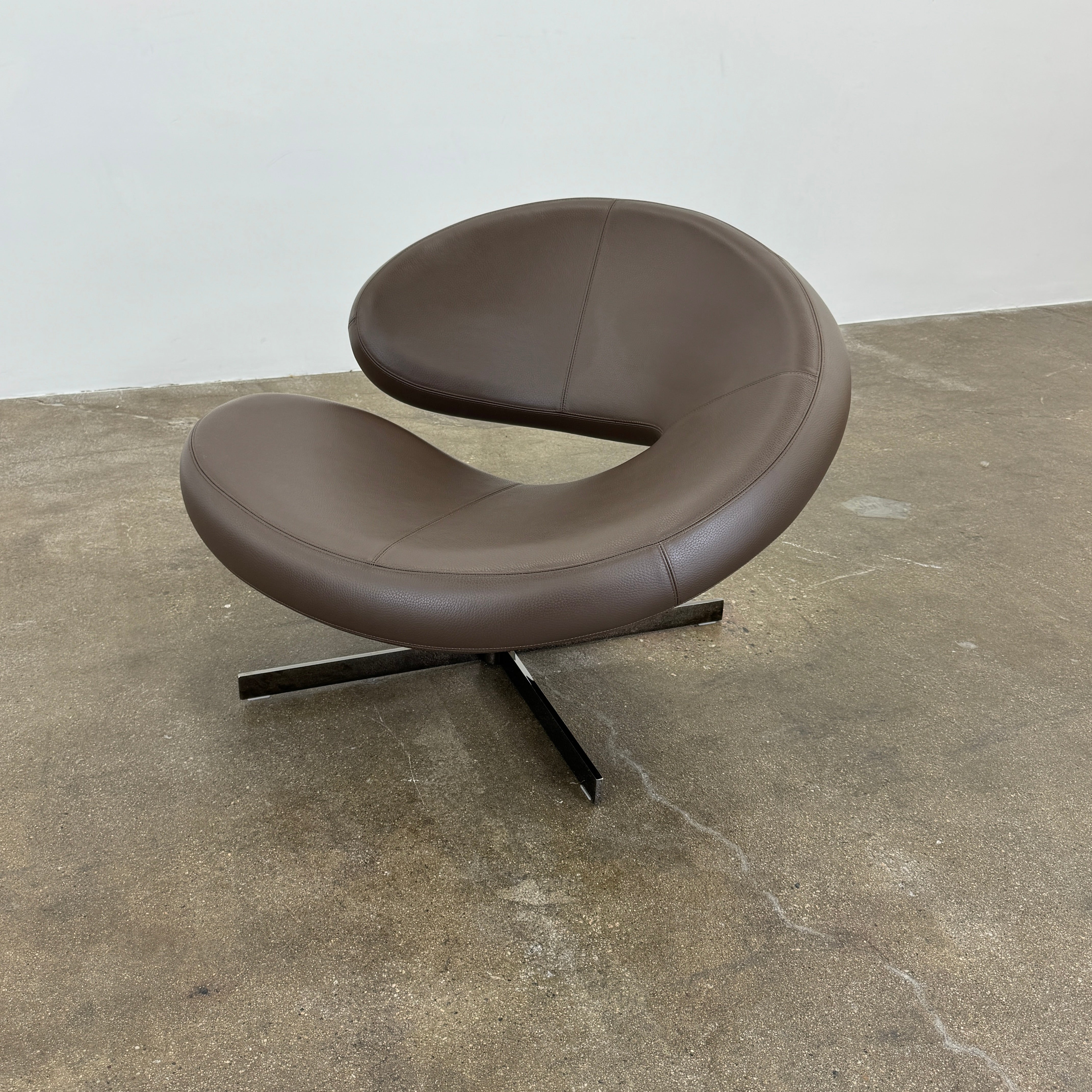 The Roche Bobois Nuage Swivel Chair is a modern, circular brown leather chair with a curved backrest and seat on a metal swivel base, set on a concrete floor against a plain white wall.