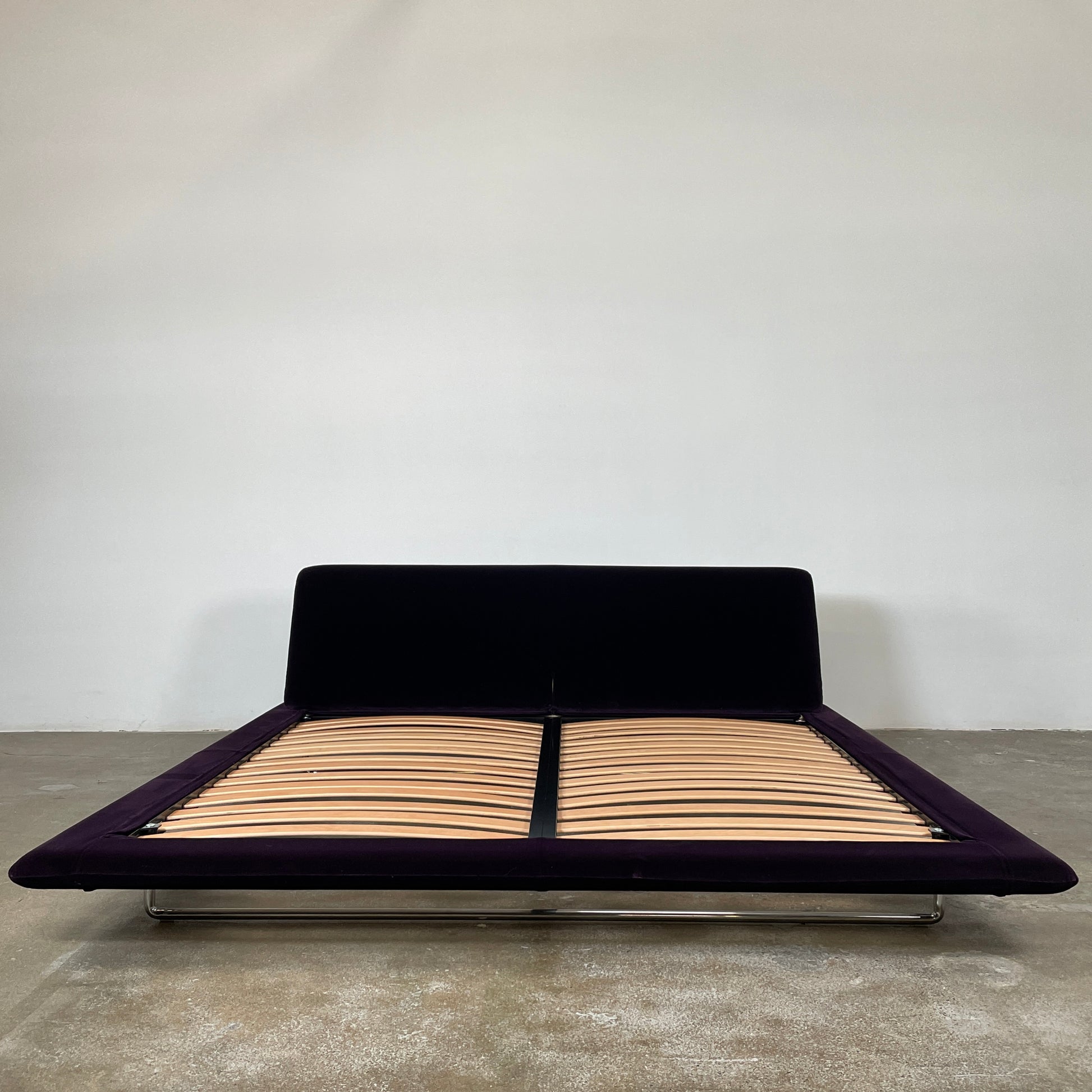 The B&B Italia Siena King Bed is a modern platform with wooden slats and an angled headboard, featuring a luxurious dark upholstered frame and sleek metal legs. Inspired by Naoto Fukasawa, it embodies understated elegance and sophistication for any contemporary bedroom setting.