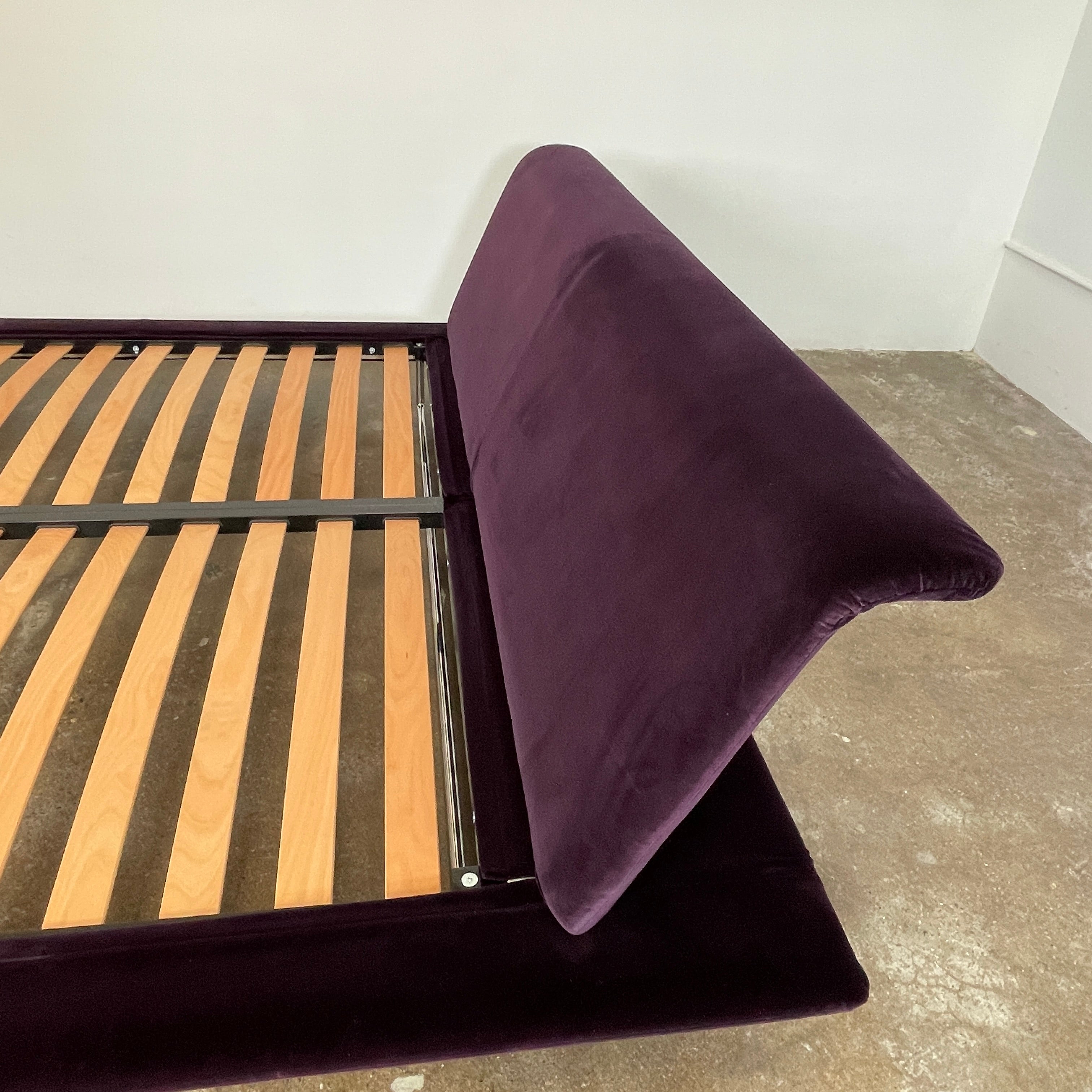 The B&B Italia Siena King Bed is a modern platform with wooden slats and an angled headboard, featuring a luxurious dark upholstered frame and sleek metal legs. Inspired by Naoto Fukasawa, it embodies understated elegance and sophistication for any contemporary bedroom setting.