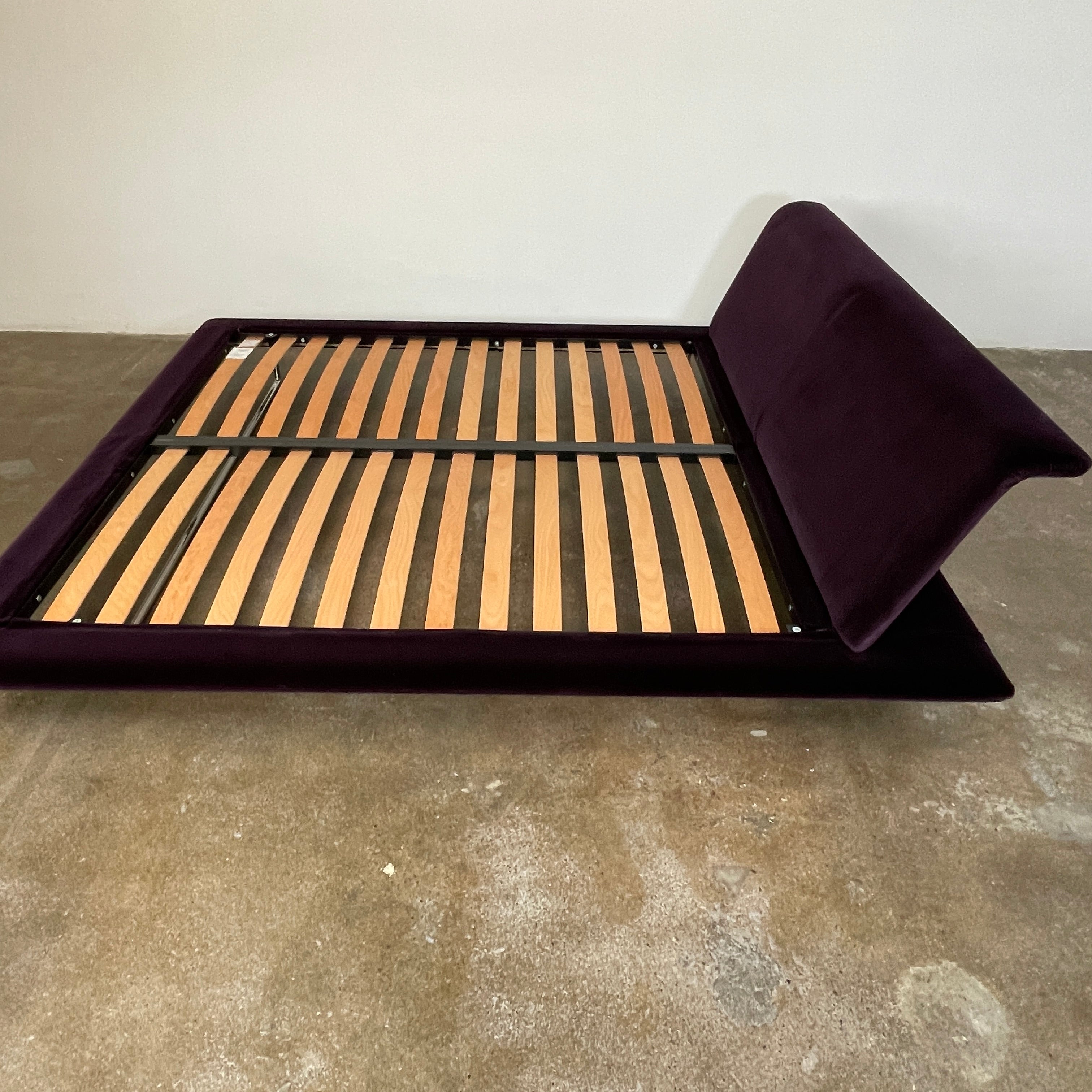 The B&B Italia Siena King Bed is a modern platform with wooden slats and an angled headboard, featuring a luxurious dark upholstered frame and sleek metal legs. Inspired by Naoto Fukasawa, it embodies understated elegance and sophistication for any contemporary bedroom setting.