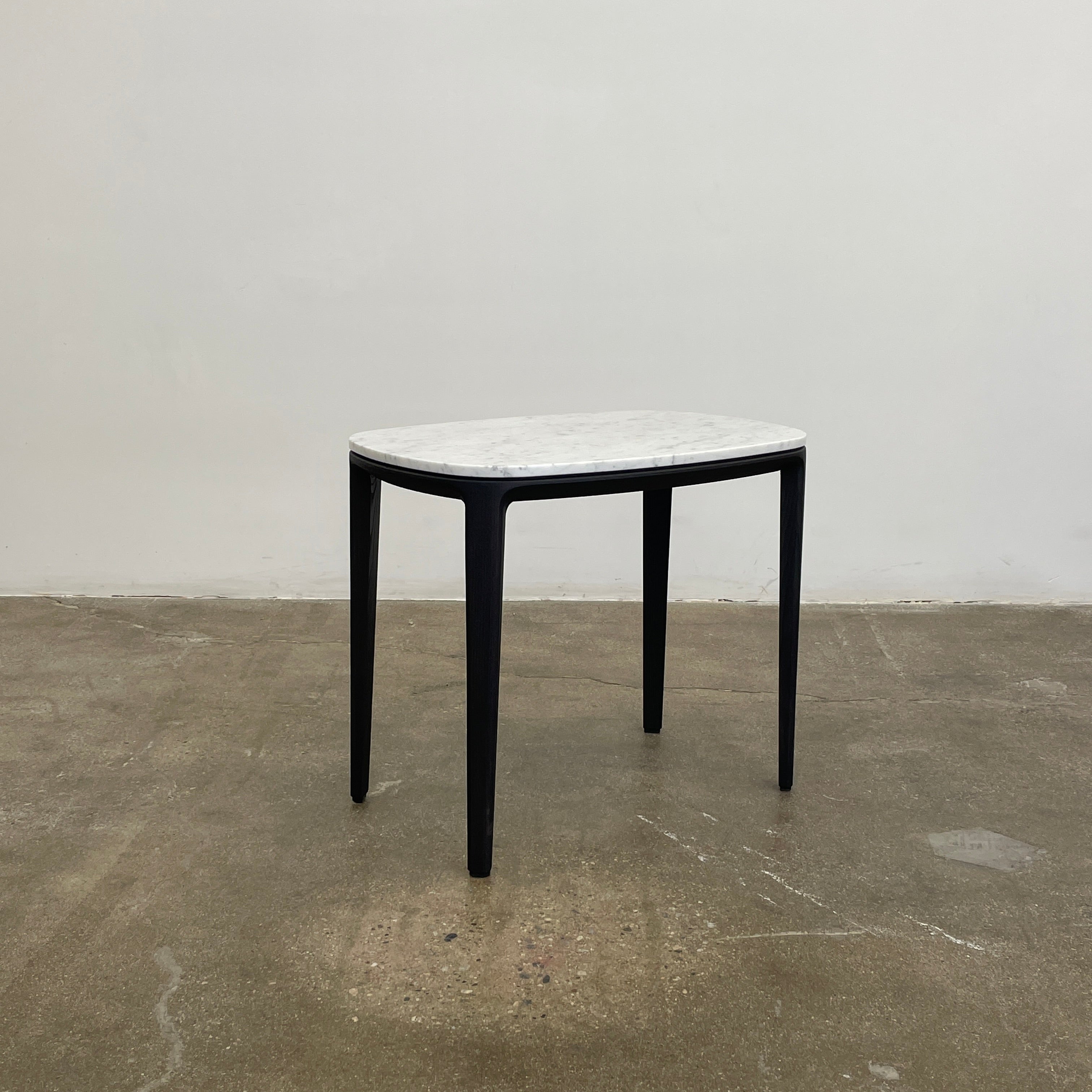 The Poliform Henry Marble Side Table, by Poliform, showcases an elegant small oval design with a luxurious Carrara marble top. It stands gracefully on four black Elm legs against a minimalist white backdrop.