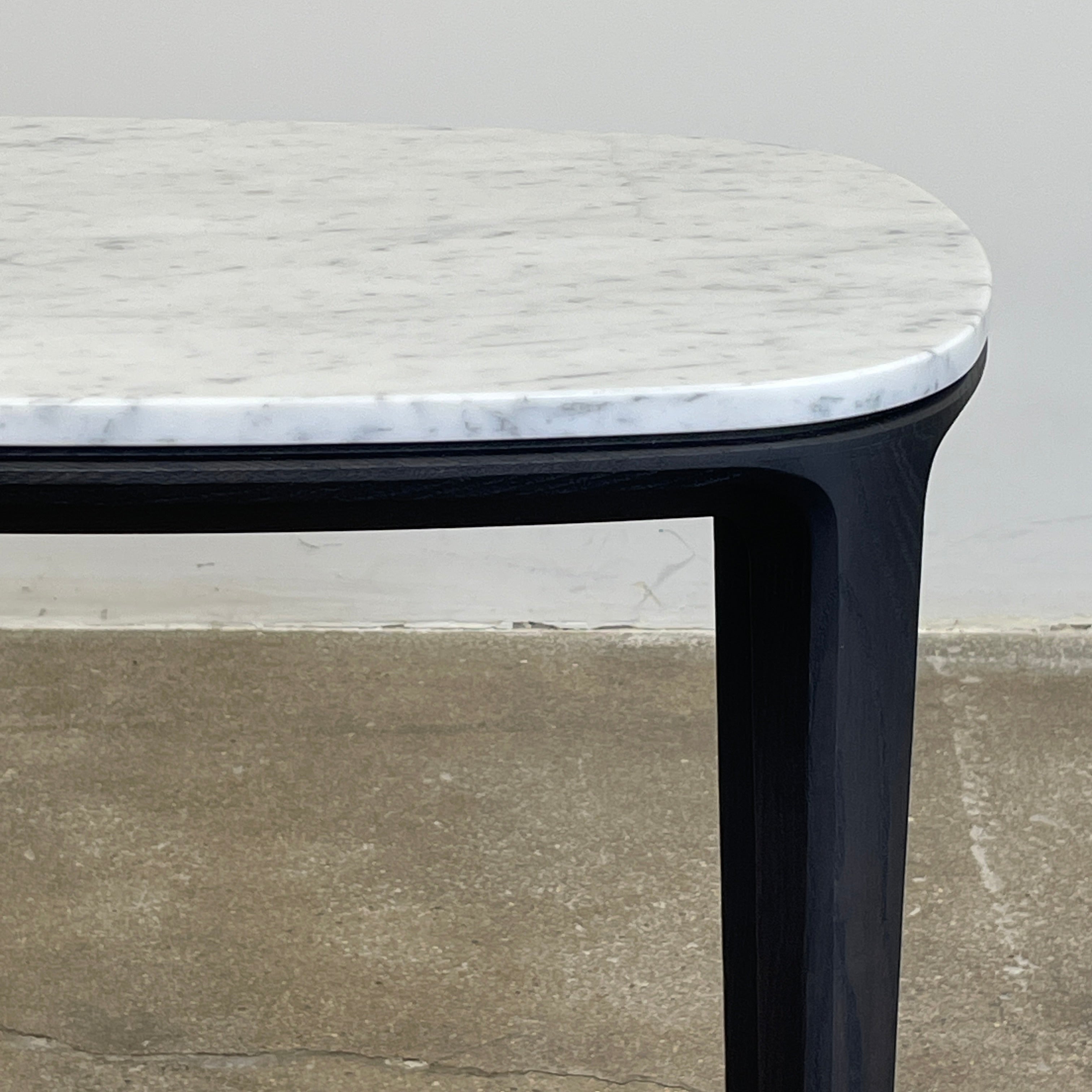 The Poliform Henry Marble Side Table, by Poliform, showcases an elegant small oval design with a luxurious Carrara marble top. It stands gracefully on four black Elm legs against a minimalist white backdrop.