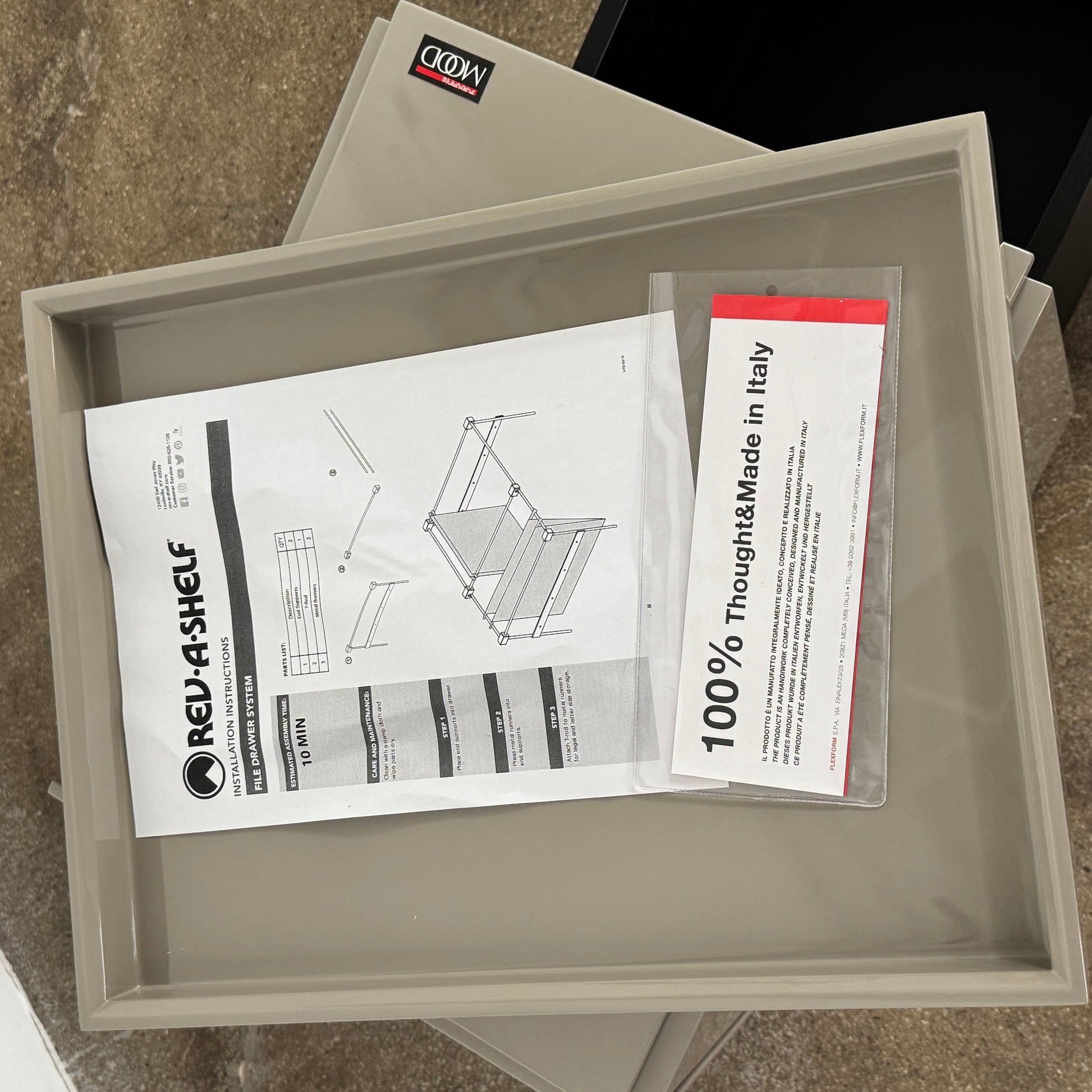 A Rev-A-Shelf instruction sheet and a "100% Thought&Made in Italy" leaflet sit on two beige trays, ideal for organizing your workspace or pairing with the Flexform Benjamin Chest of Drawers With File Insert.