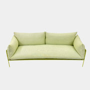 The light green Paola Lenti Kaba Outdoor Sofa by Modern Resale, featuring a metal frame, functions as a stylish two-seater and is shown against a plain white wall on a brown concrete floor.