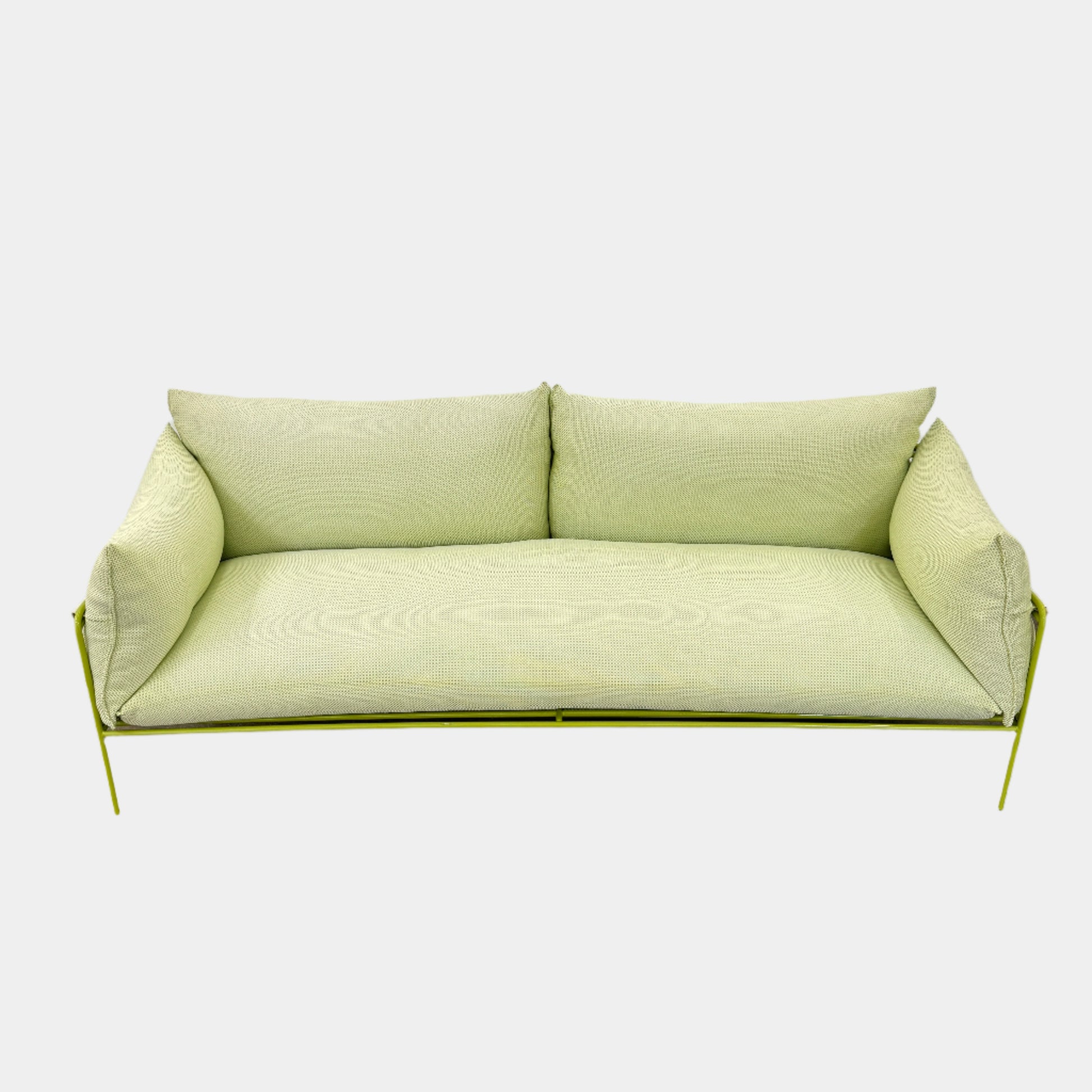 The light green Paola Lenti Kaba Outdoor Sofa by Modern Resale, featuring a metal frame, functions as a stylish two-seater and is shown against a plain white wall on a brown concrete floor.