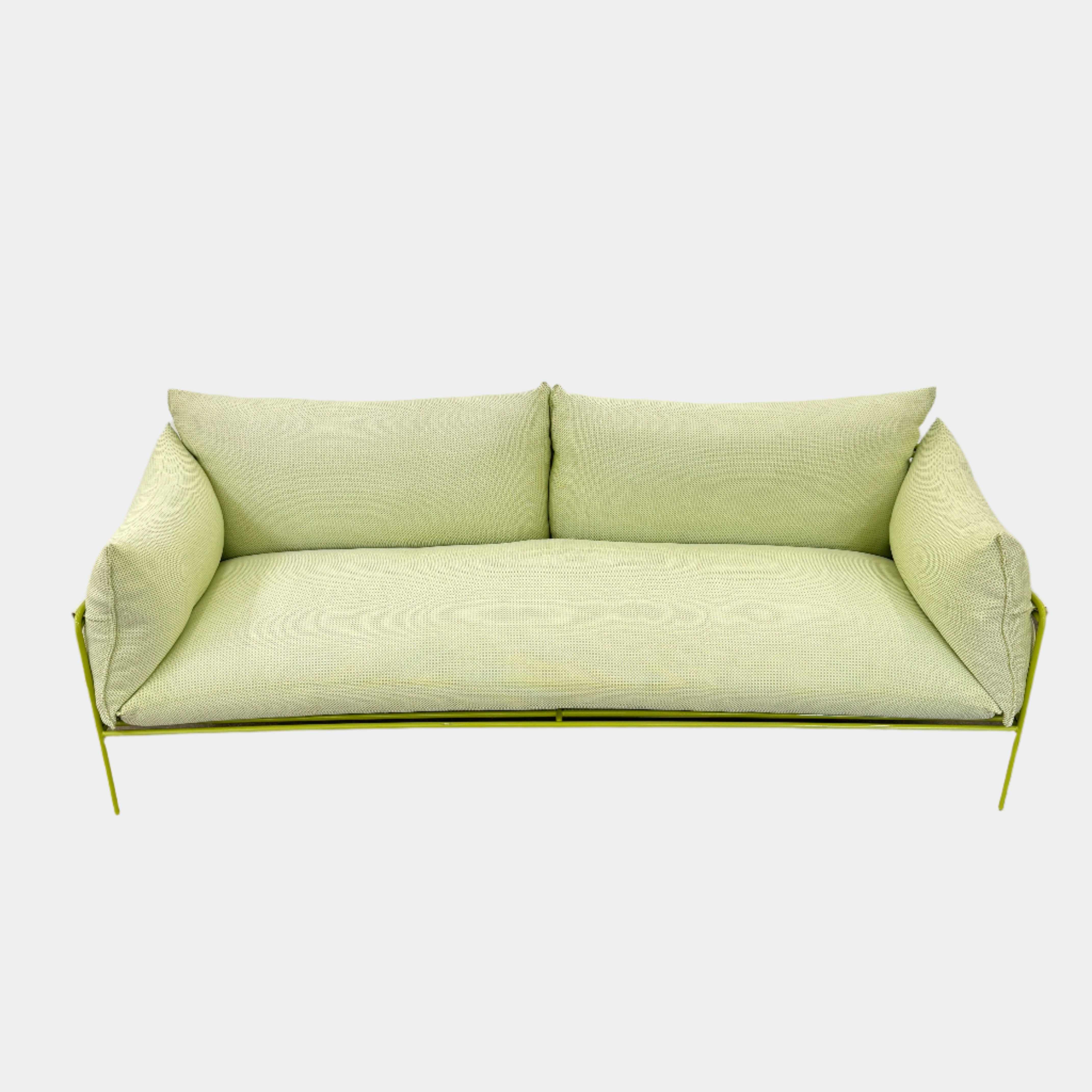 The light green Paola Lenti Kaba Outdoor Sofa by Modern Resale, featuring a metal frame, functions as a stylish two-seater and is shown against a plain white wall on a brown concrete floor.