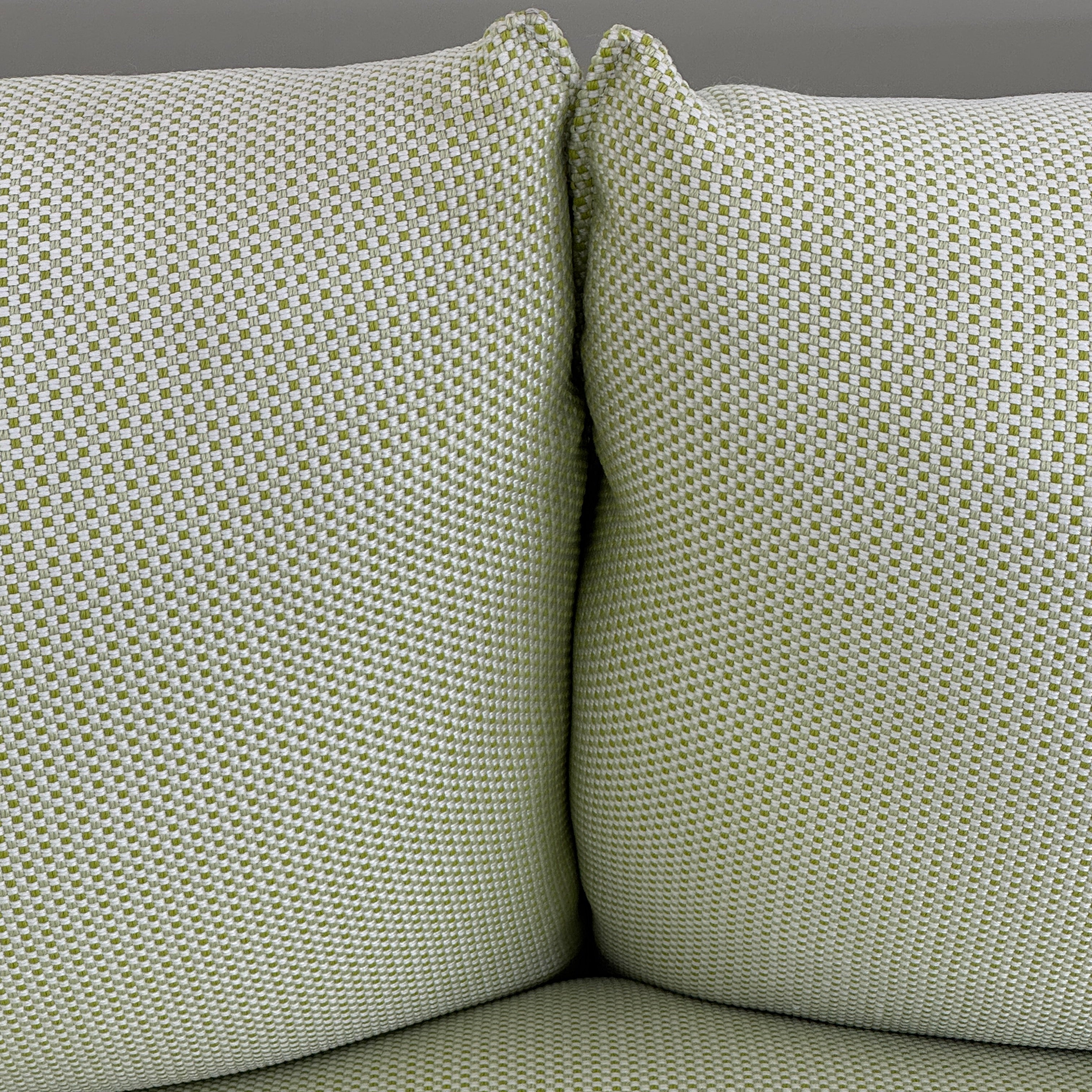 The light green Paola Lenti Kaba Outdoor Sofa by Modern Resale, featuring a metal frame, functions as a stylish two-seater and is shown against a plain white wall on a brown concrete floor.