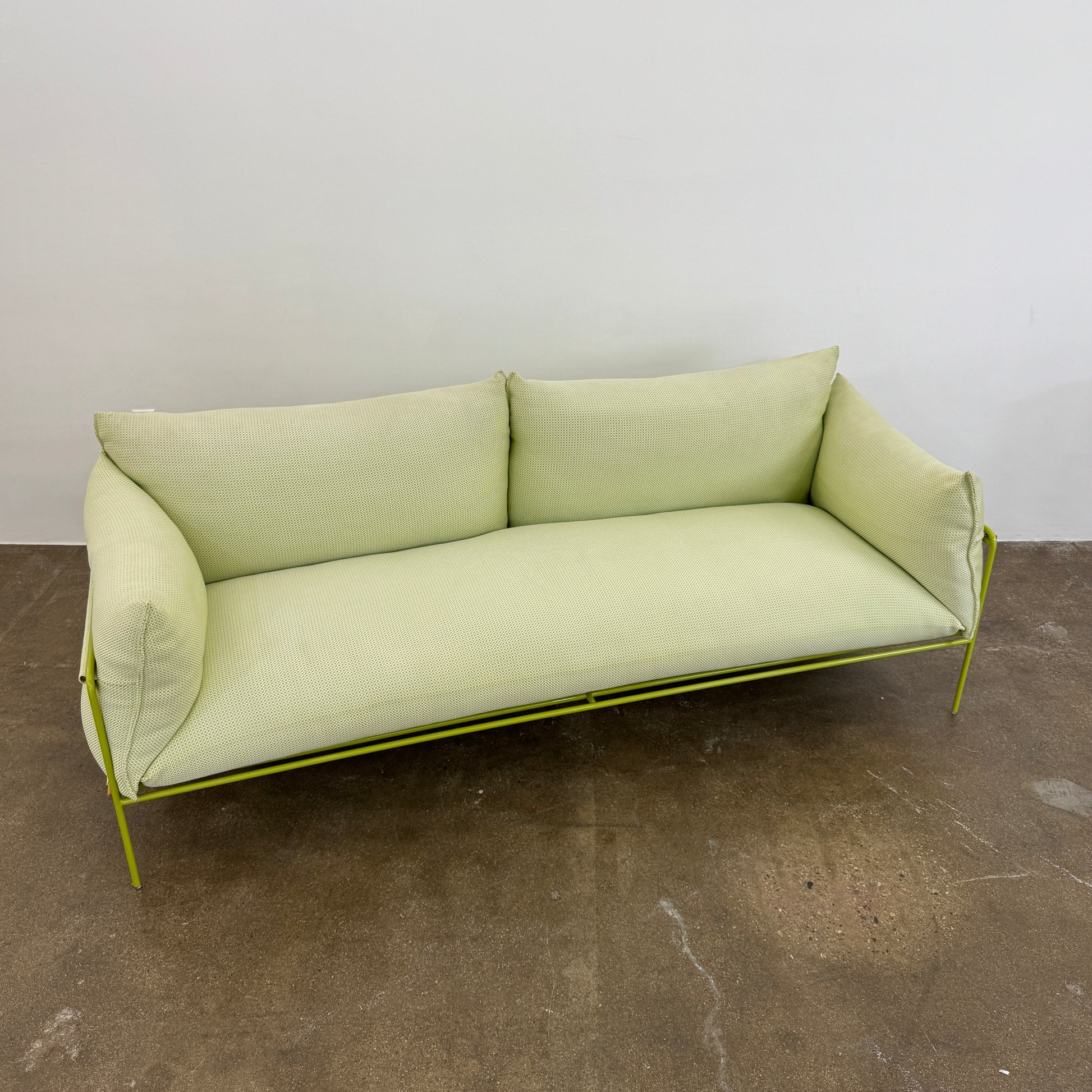 The light green Paola Lenti Kaba Outdoor Sofa by Modern Resale, featuring a metal frame, functions as a stylish two-seater and is shown against a plain white wall on a brown concrete floor.