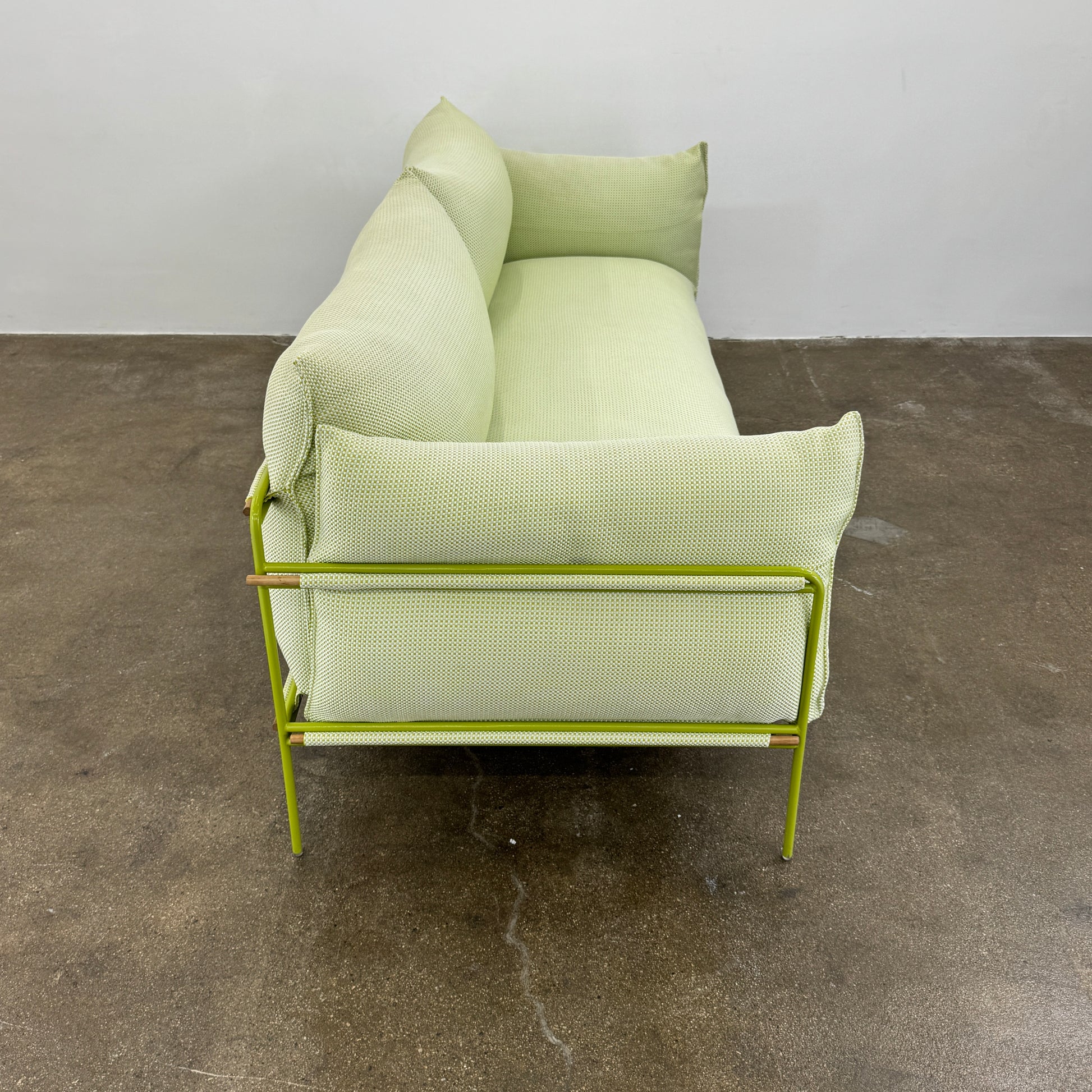 The light green Paola Lenti Kaba Outdoor Sofa by Modern Resale, featuring a metal frame, functions as a stylish two-seater and is shown against a plain white wall on a brown concrete floor.