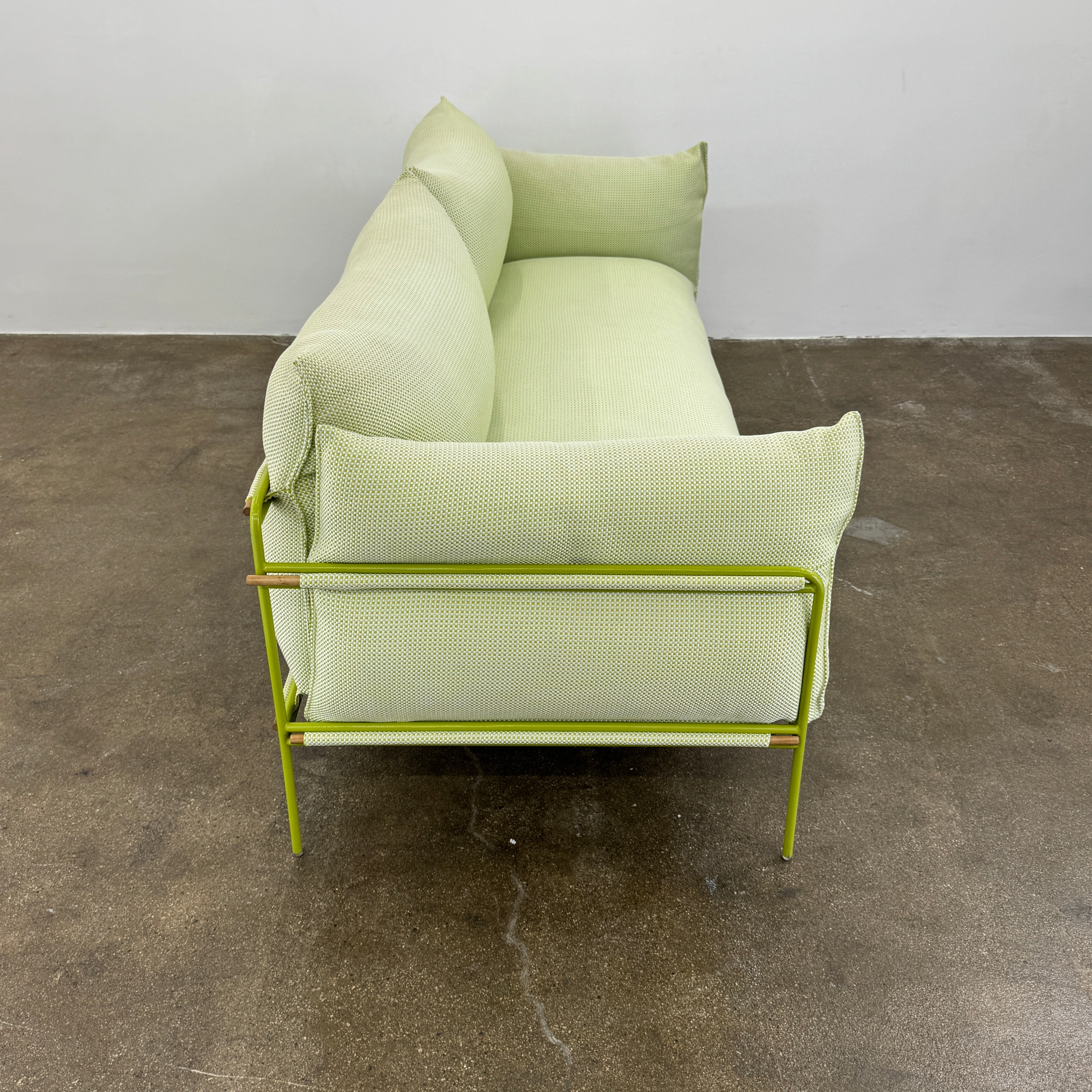 The light green Paola Lenti Kaba Outdoor Sofa by Modern Resale, featuring a metal frame, functions as a stylish two-seater and is shown against a plain white wall on a brown concrete floor.