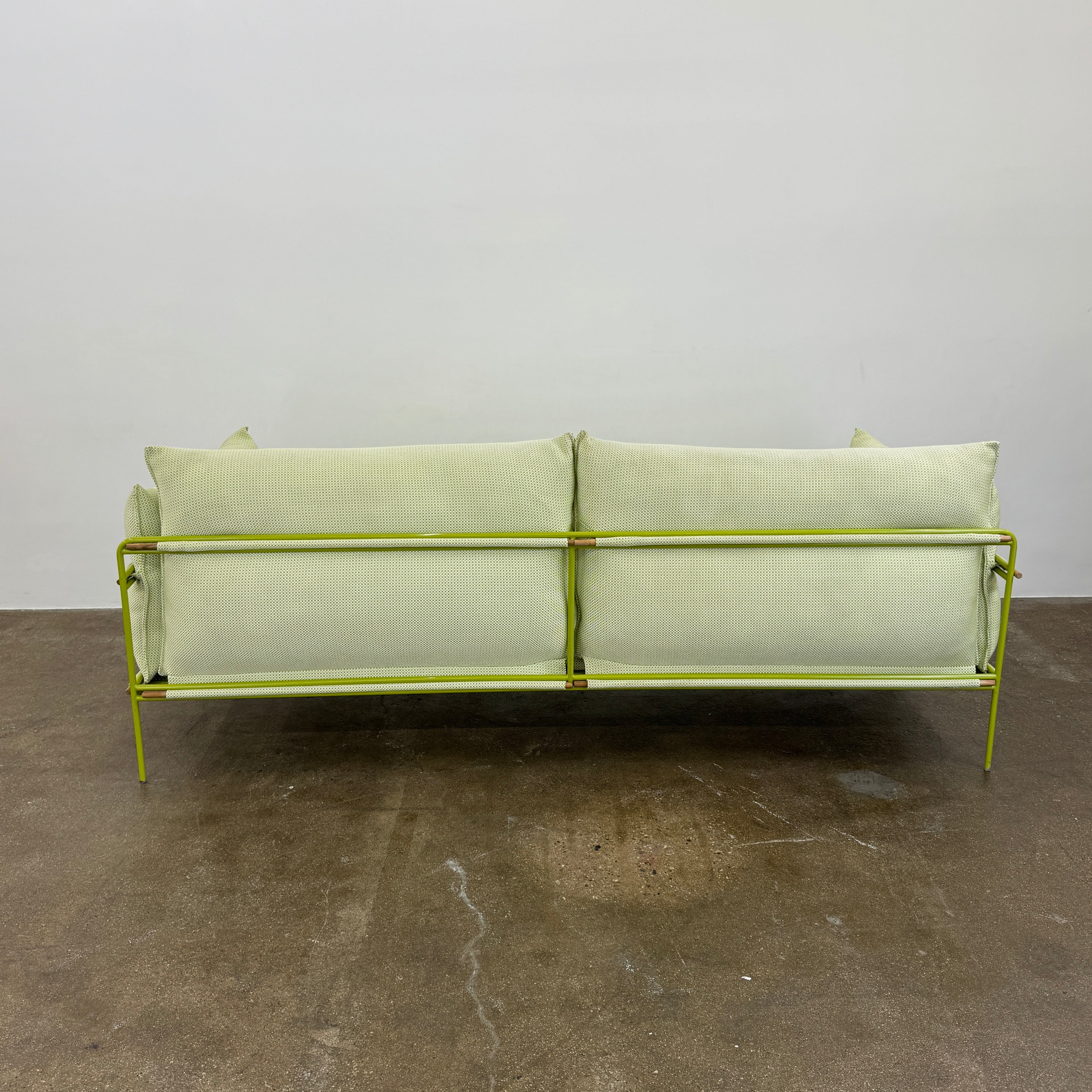 The light green Paola Lenti Kaba Outdoor Sofa by Modern Resale, featuring a metal frame, functions as a stylish two-seater and is shown against a plain white wall on a brown concrete floor.