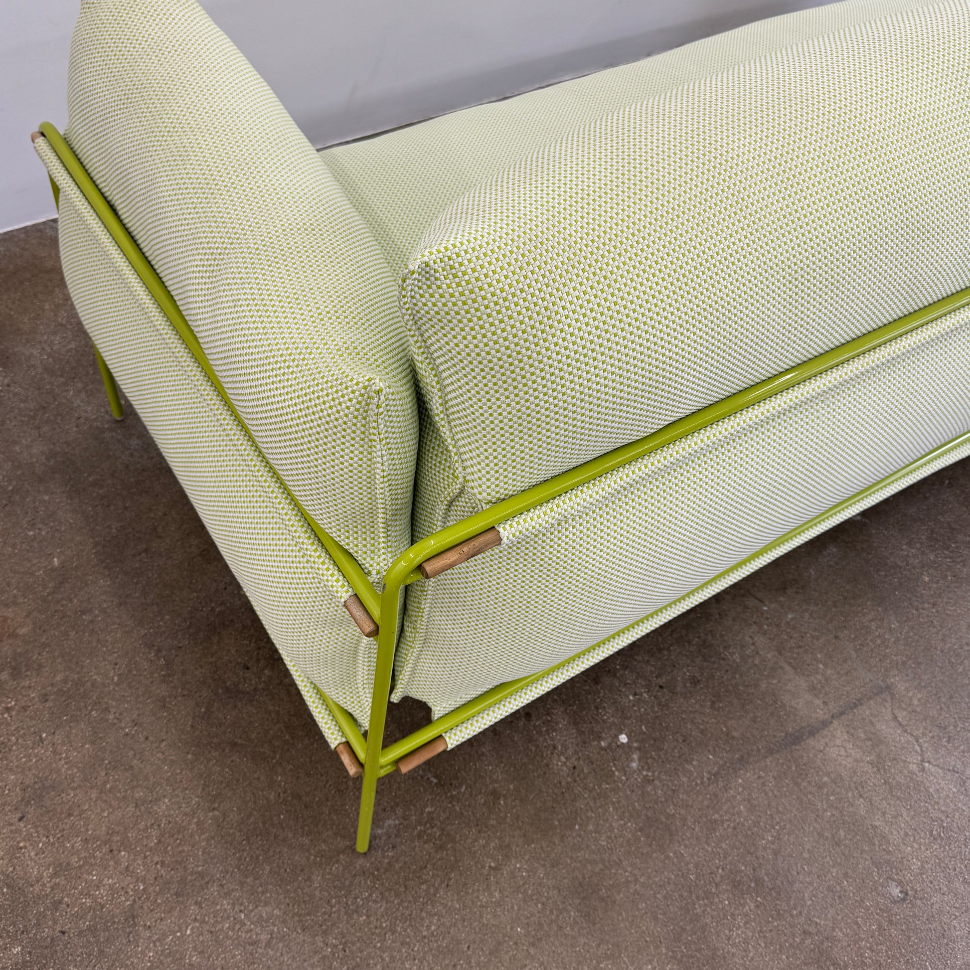 The light green Paola Lenti Kaba Outdoor Sofa by Modern Resale, featuring a metal frame, functions as a stylish two-seater and is shown against a plain white wall on a brown concrete floor.