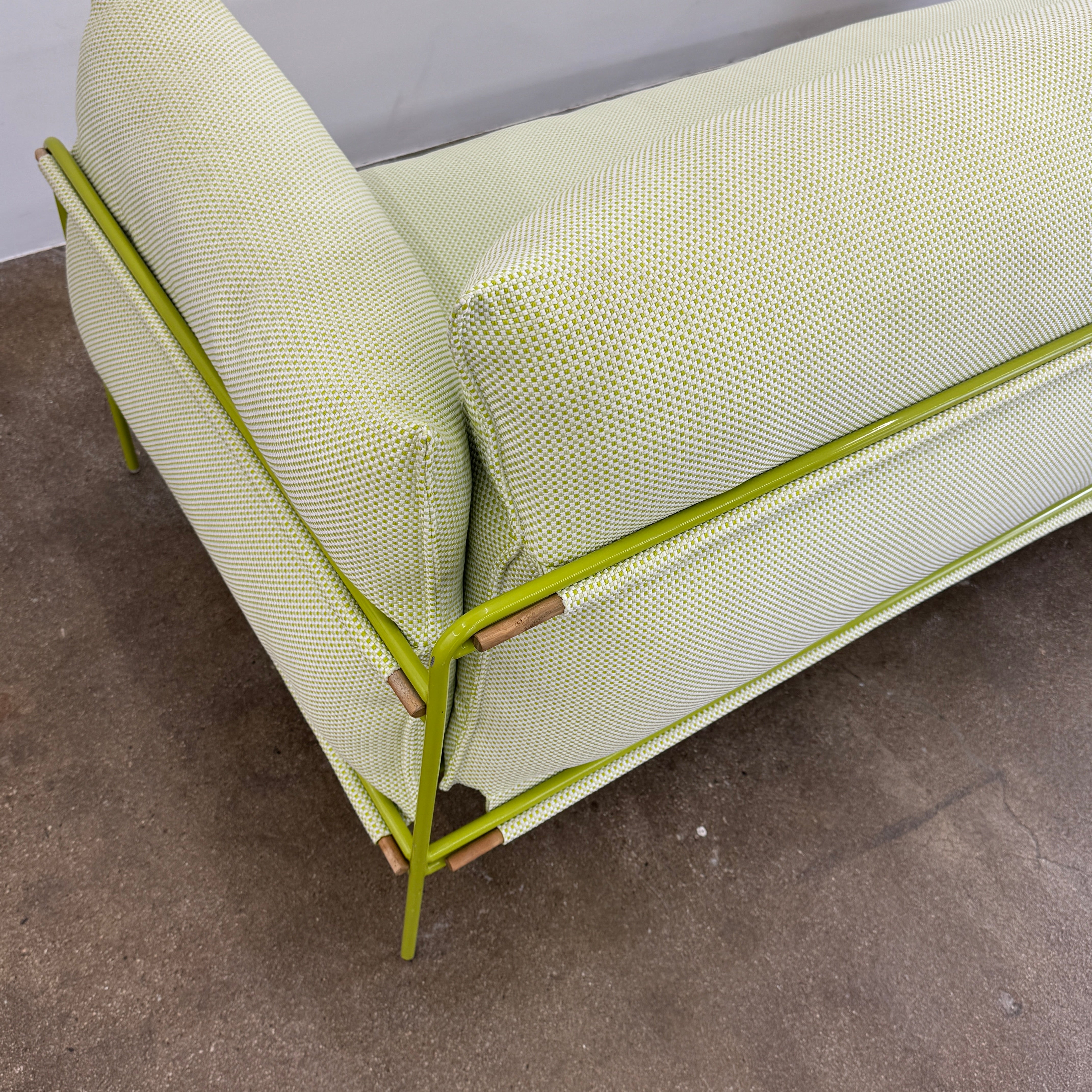 The light green Paola Lenti Kaba Outdoor Sofa by Modern Resale, featuring a metal frame, functions as a stylish two-seater and is shown against a plain white wall on a brown concrete floor.