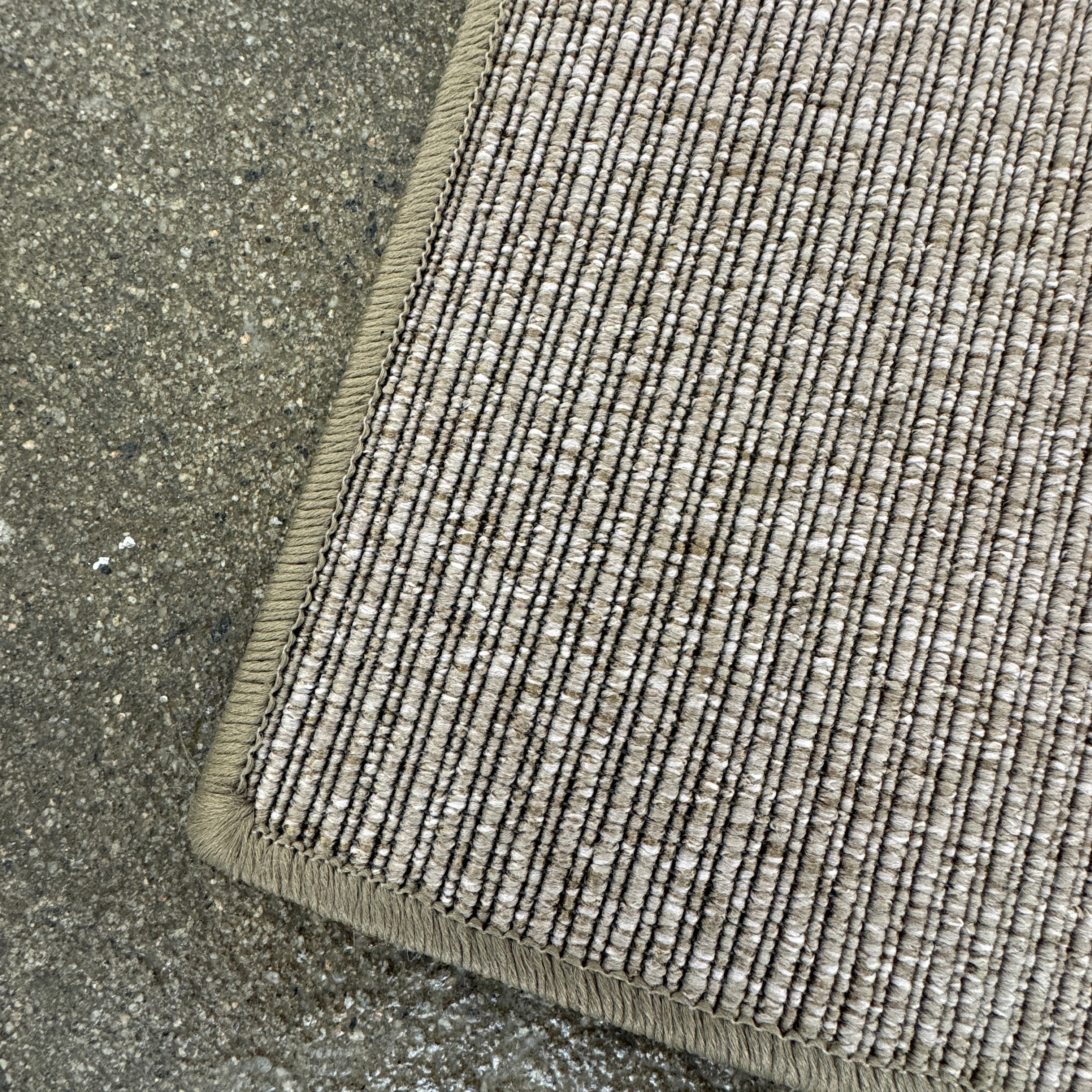 A beige Flexform Paride Rug with stitched edges rests on a polished concrete floor.