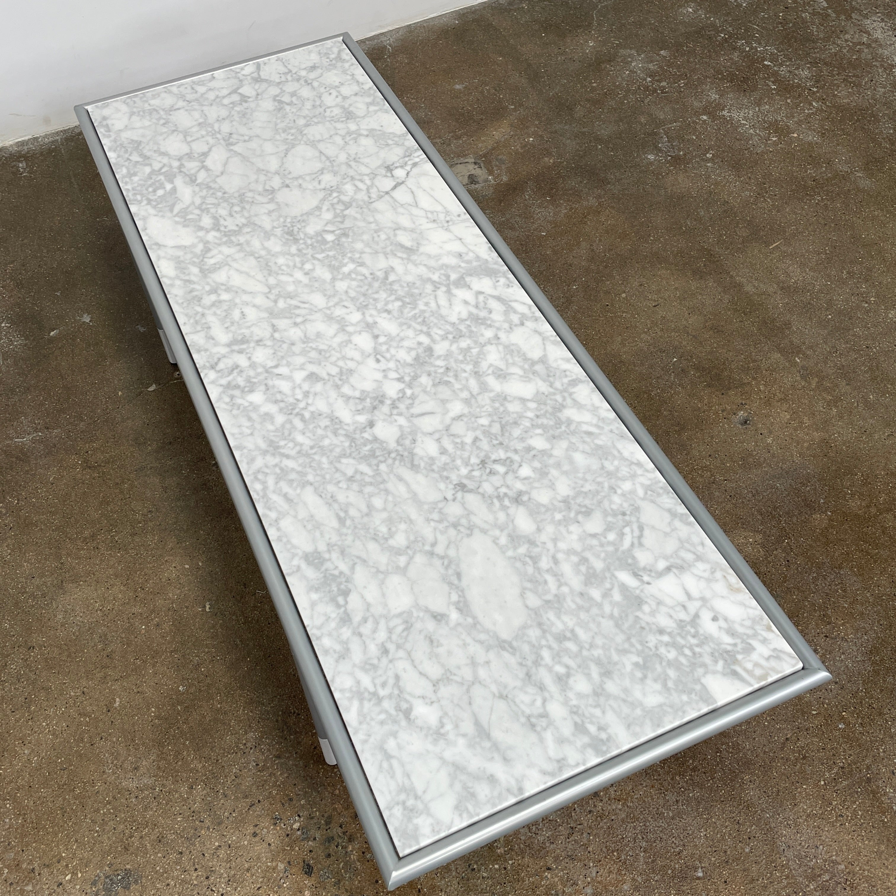 The Artelano Carrara Marble Coffee Table boasts a rectangular marble top with sleek metal legs and a central wood tray, exuding the Parisian elegance of the Artelano brand.