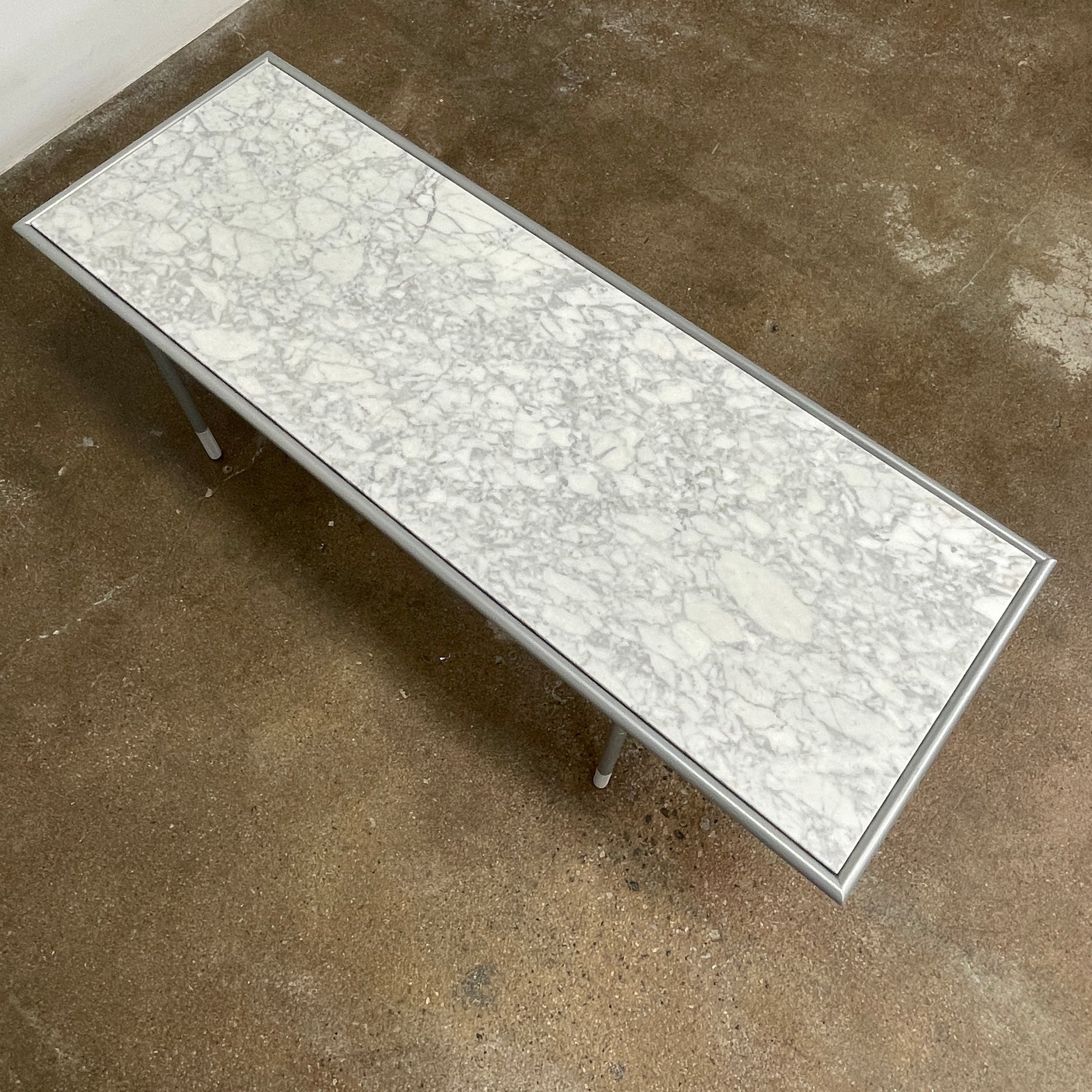 The Artelano Carrara Marble Coffee Table boasts a rectangular marble top with sleek metal legs and a central wood tray, exuding the Parisian elegance of the Artelano brand.