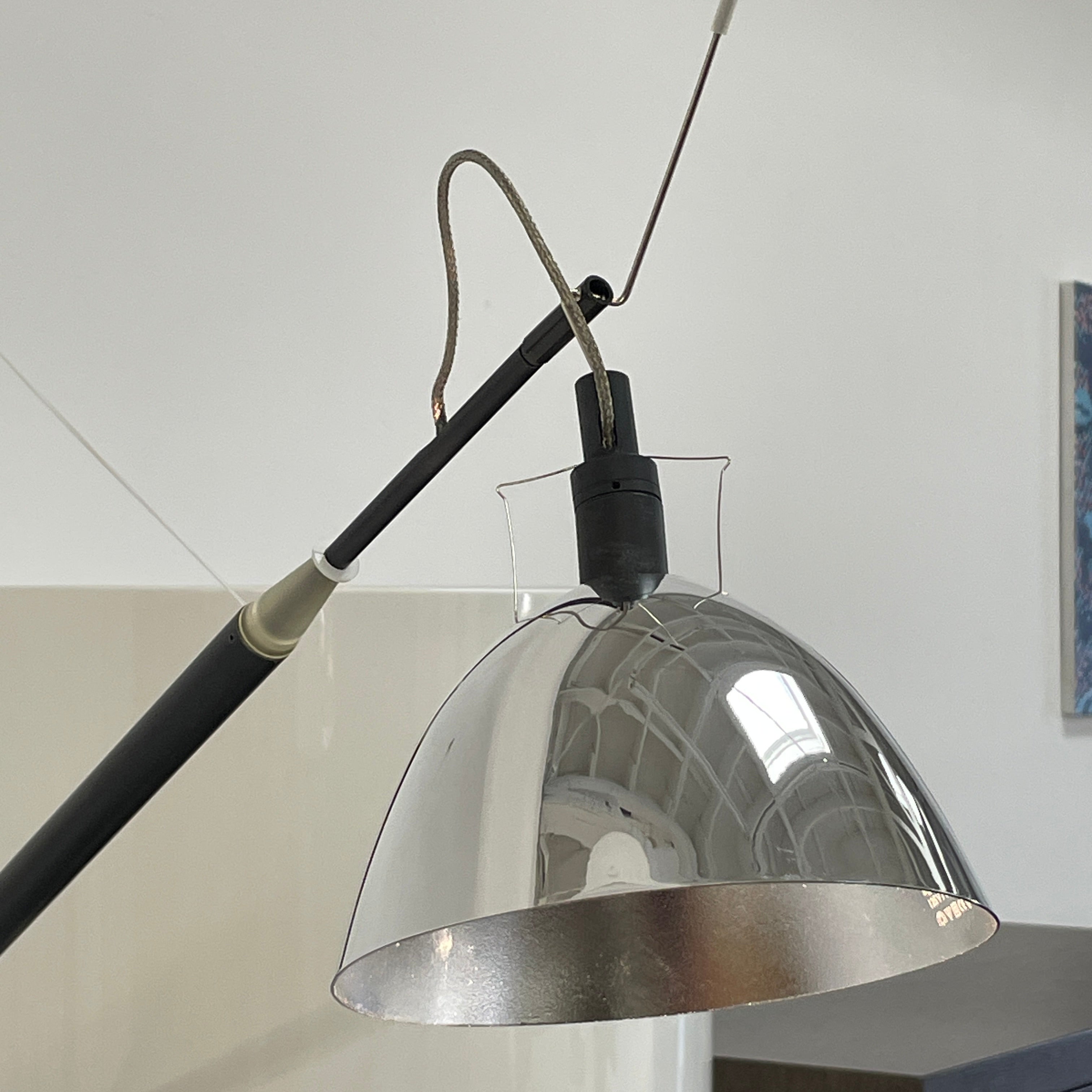 The Ingo Maurer Max Mover Wall Light by Ingo Maurer is a contemporary fixture with an adjustable telescopic rod, sleek black arm, and aluminum reflector in a dome-shaped shade set against a light grey backdrop.
