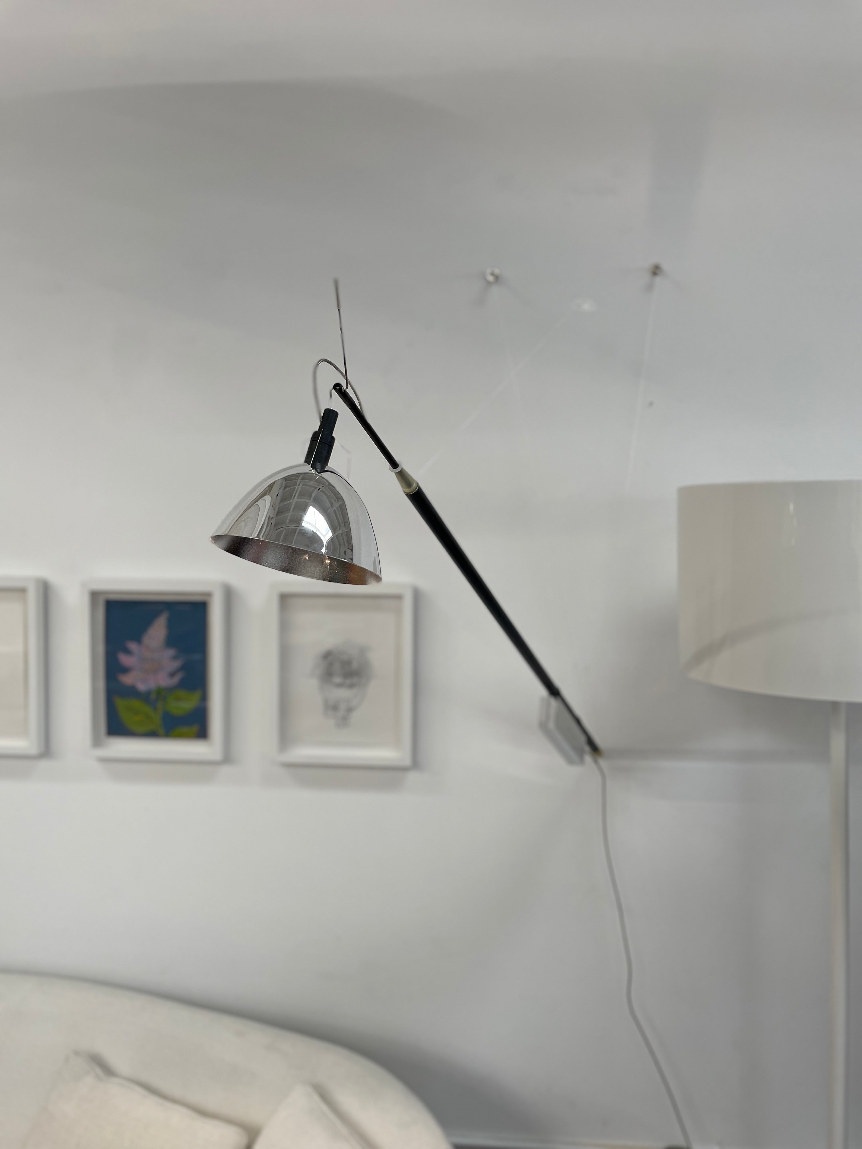 The Ingo Maurer Max Mover Wall Light by Ingo Maurer is a contemporary fixture with an adjustable telescopic rod, sleek black arm, and aluminum reflector in a dome-shaped shade set against a light grey backdrop.