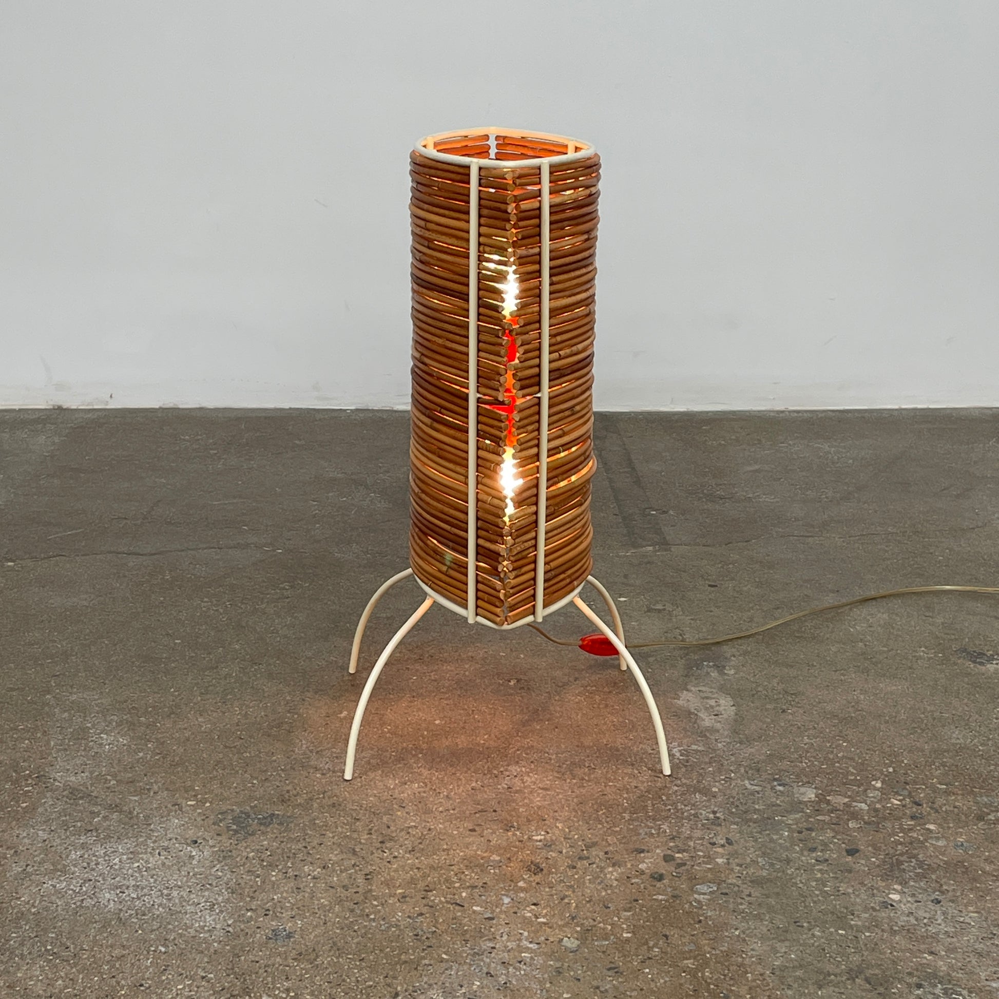 The Vintage Fontana Arte Bambú Table Light by Fontana Arte showcases a woven rattan circular shade atop a sleek white metal tripod base, making it an elegant addition to a wooden table. This lamp enhances your home's interior lighting with its natural charm and unique design.