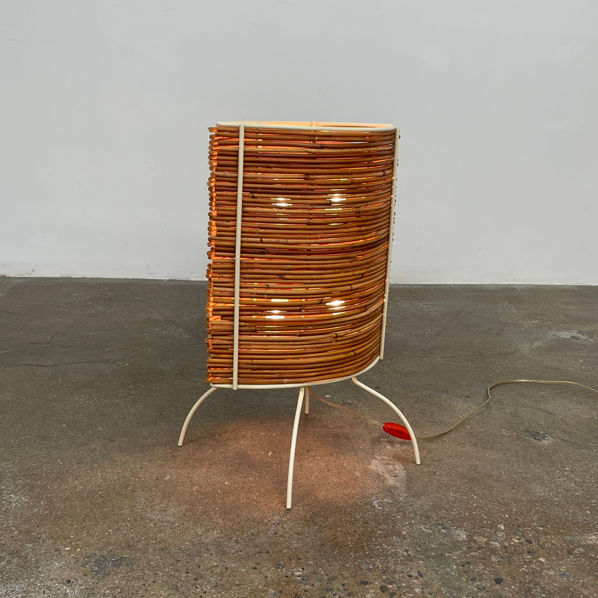 The Vintage Fontana Arte Bambú Table Light by Fontana Arte showcases a woven rattan circular shade atop a sleek white metal tripod base, making it an elegant addition to a wooden table. This lamp enhances your home's interior lighting with its natural charm and unique design.