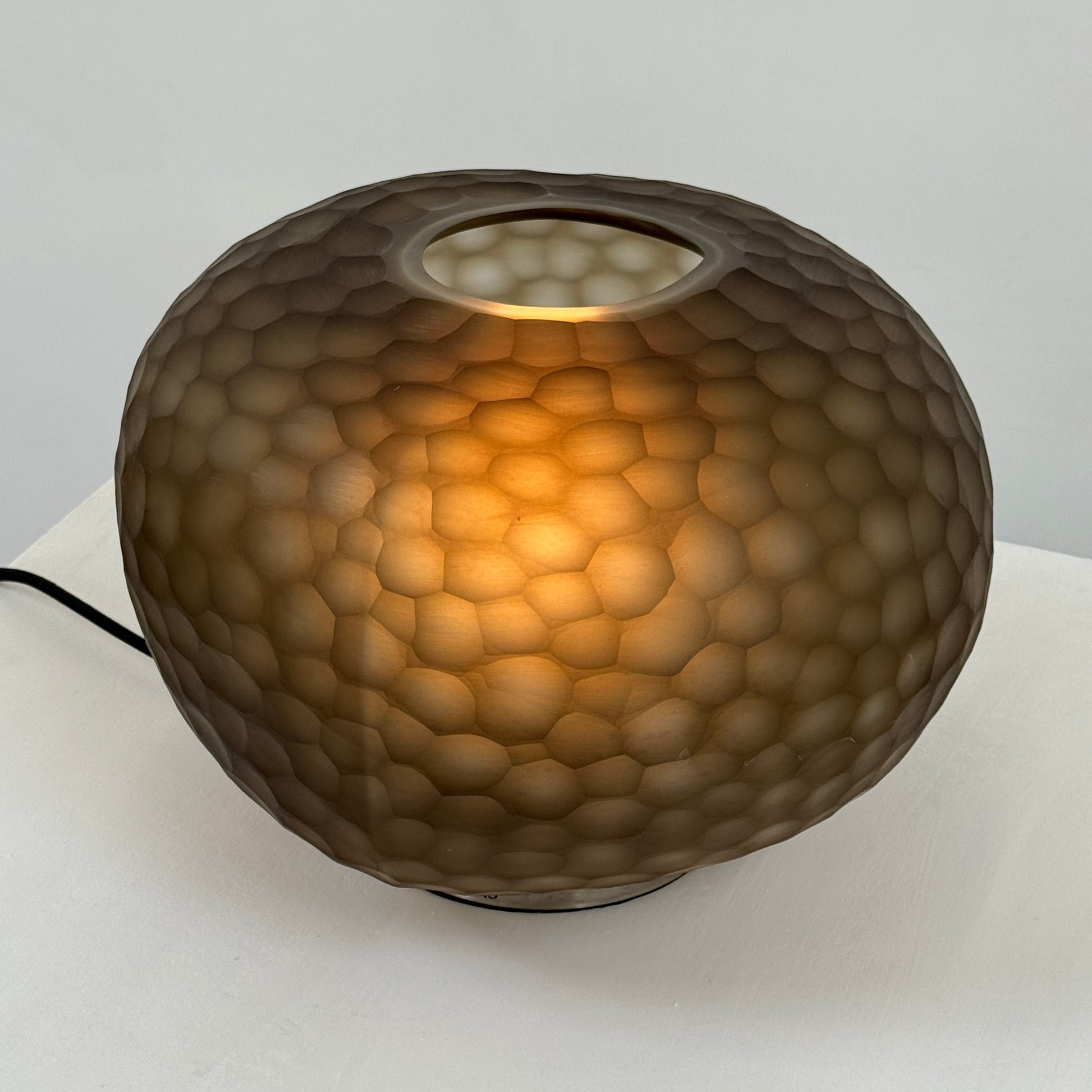 The Guax Erbse 2 Table Lamp by Guax features a mouth-blown, honeycomb-patterned glass shade on a round metal base, with an exposed black power cord for a touch of modern elegance.