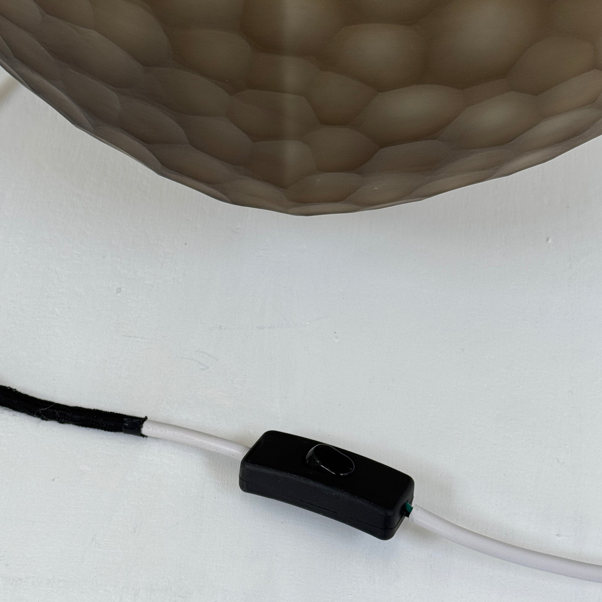 The Guax Erbse 2 Table Lamp by Guax features a mouth-blown, honeycomb-patterned glass shade on a round metal base, with an exposed black power cord for a touch of modern elegance.