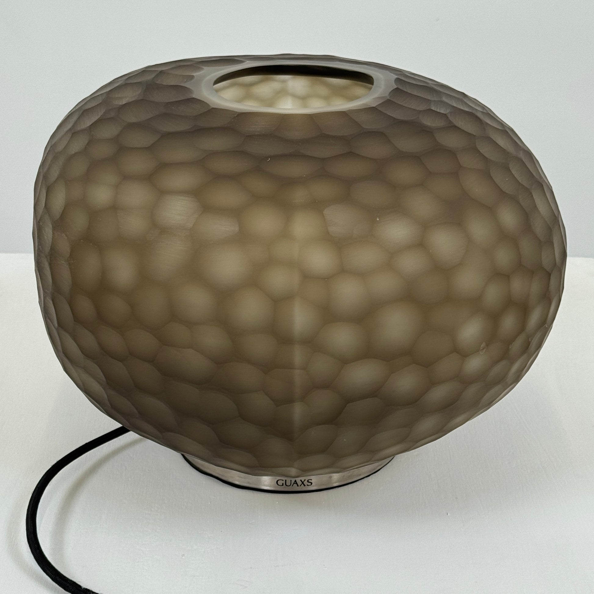 The Guax Erbse 2 Table Lamp by Guax features a mouth-blown, honeycomb-patterned glass shade on a round metal base, with an exposed black power cord for a touch of modern elegance.