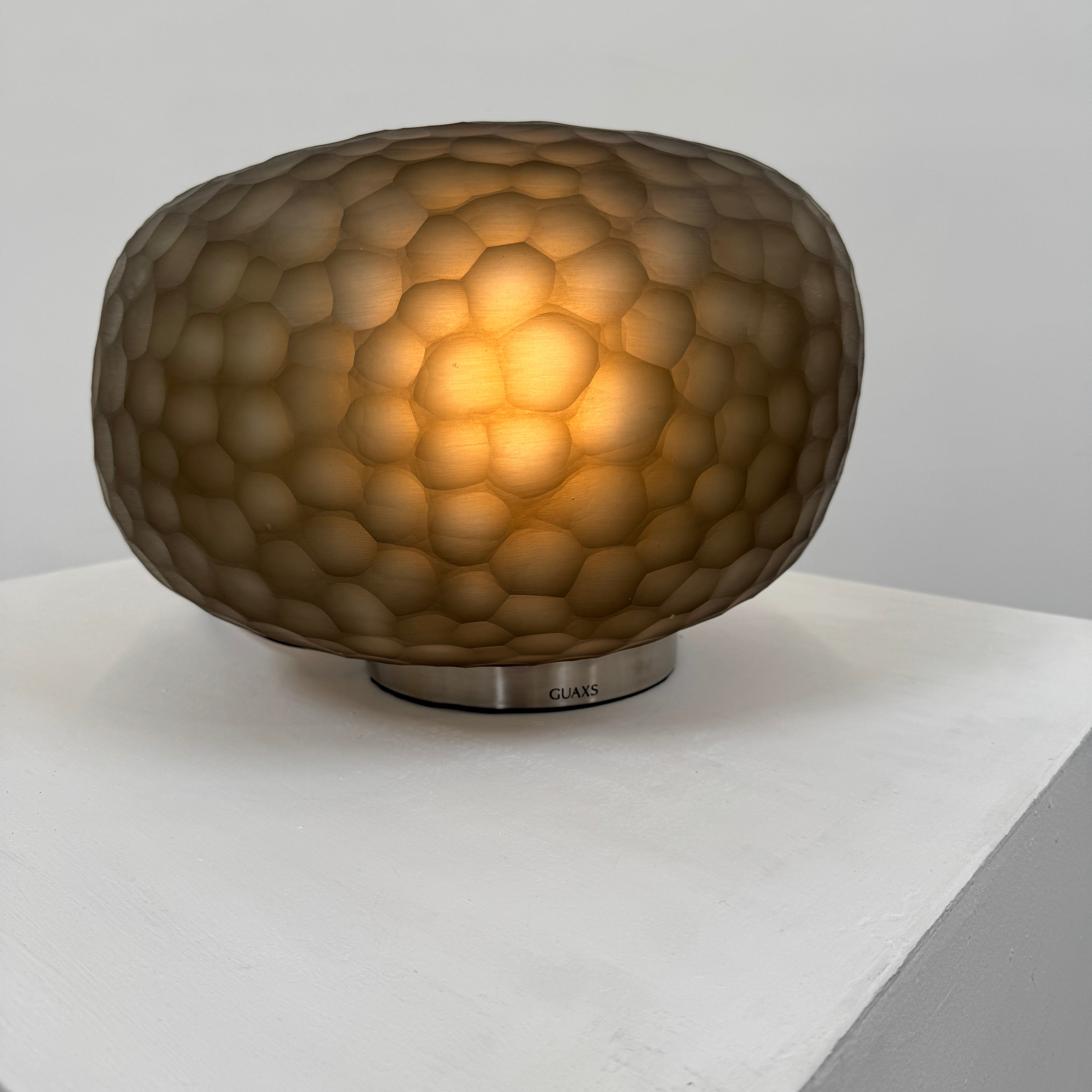 The Guax Erbse 2 Table Lamp by Guax features a mouth-blown, honeycomb-patterned glass shade on a round metal base, with an exposed black power cord for a touch of modern elegance.