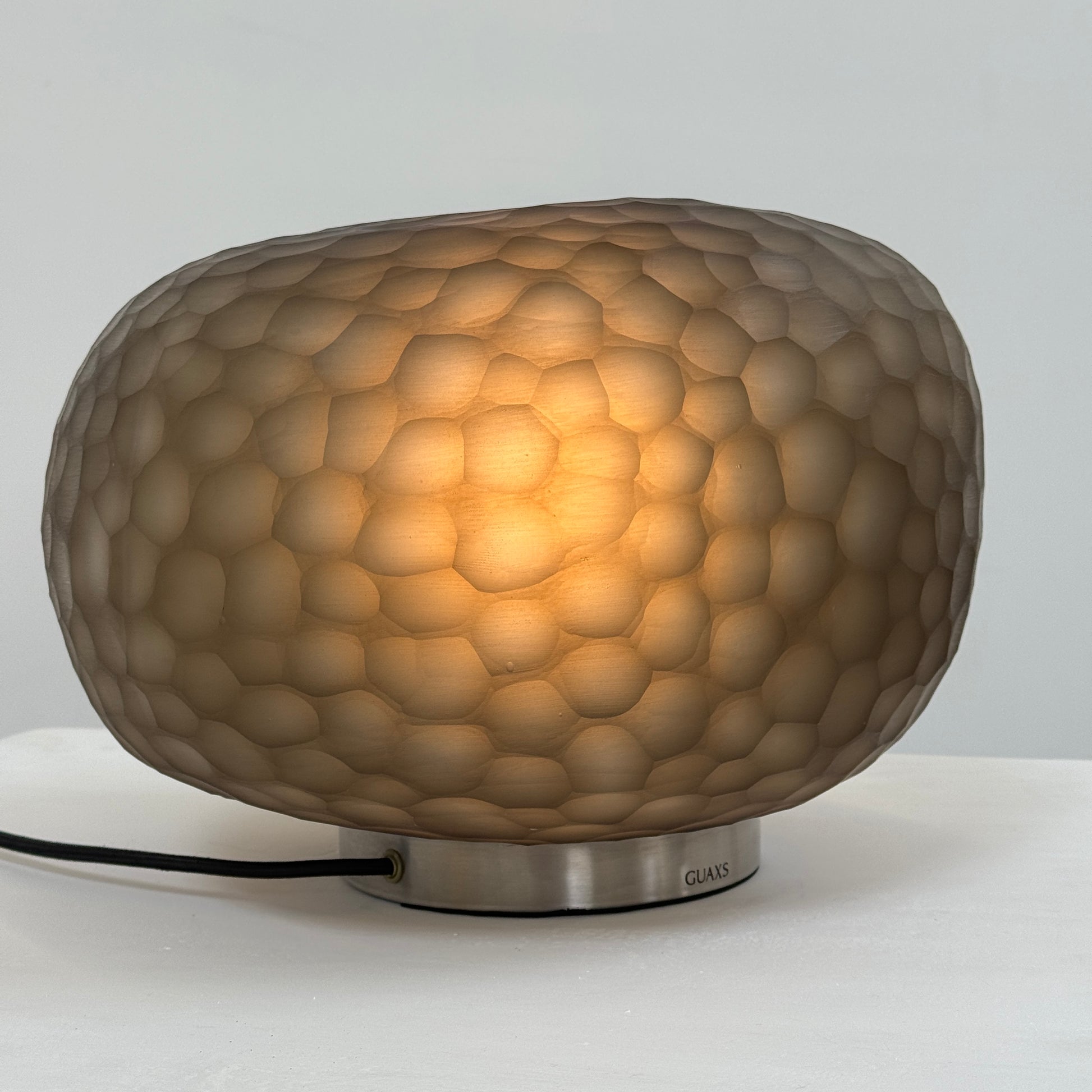 The Guax Erbse 2 Table Lamp by Guax features a mouth-blown, honeycomb-patterned glass shade on a round metal base, with an exposed black power cord for a touch of modern elegance.