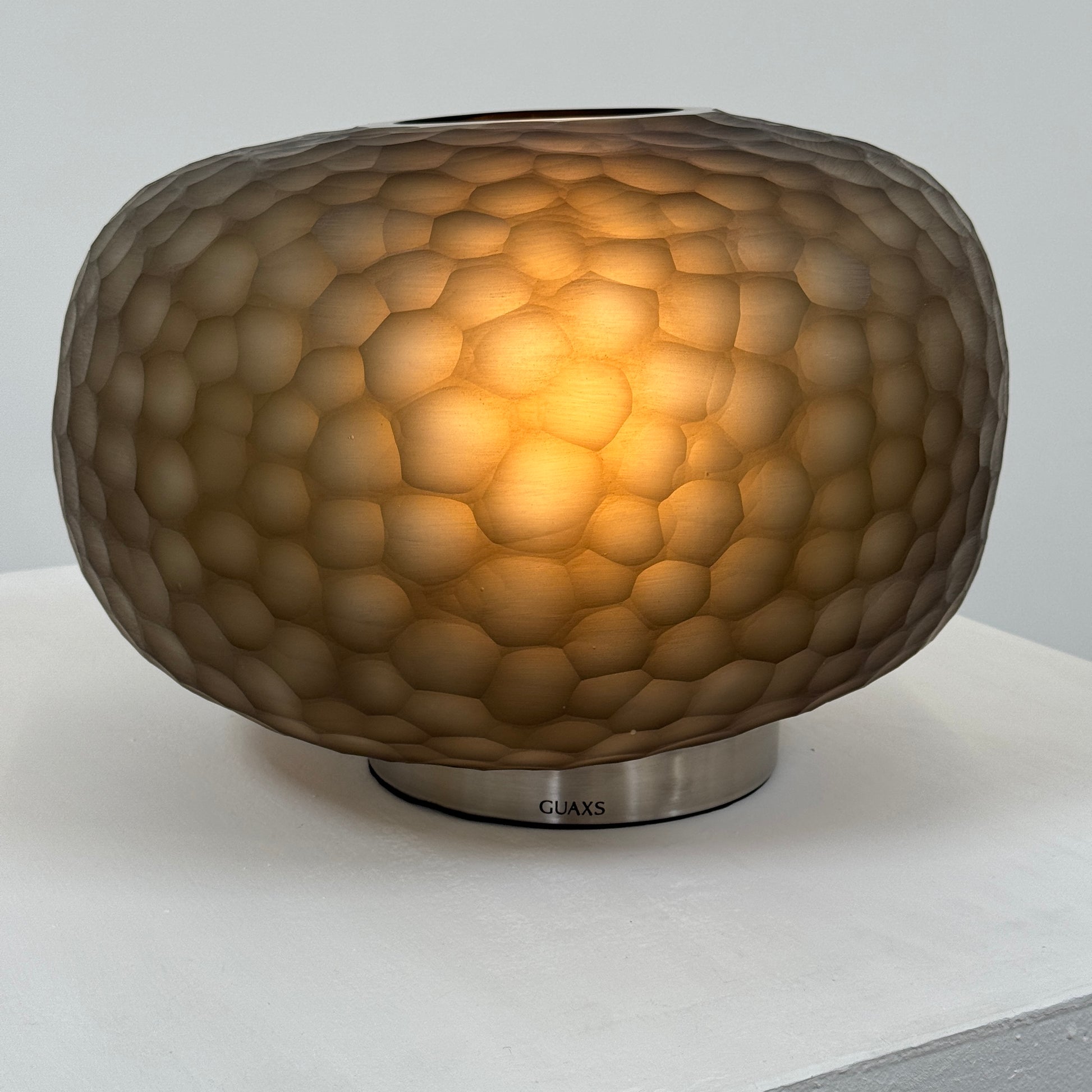 The Guax Erbse 2 Table Lamp by Guax features a mouth-blown, honeycomb-patterned glass shade on a round metal base, with an exposed black power cord for a touch of modern elegance.