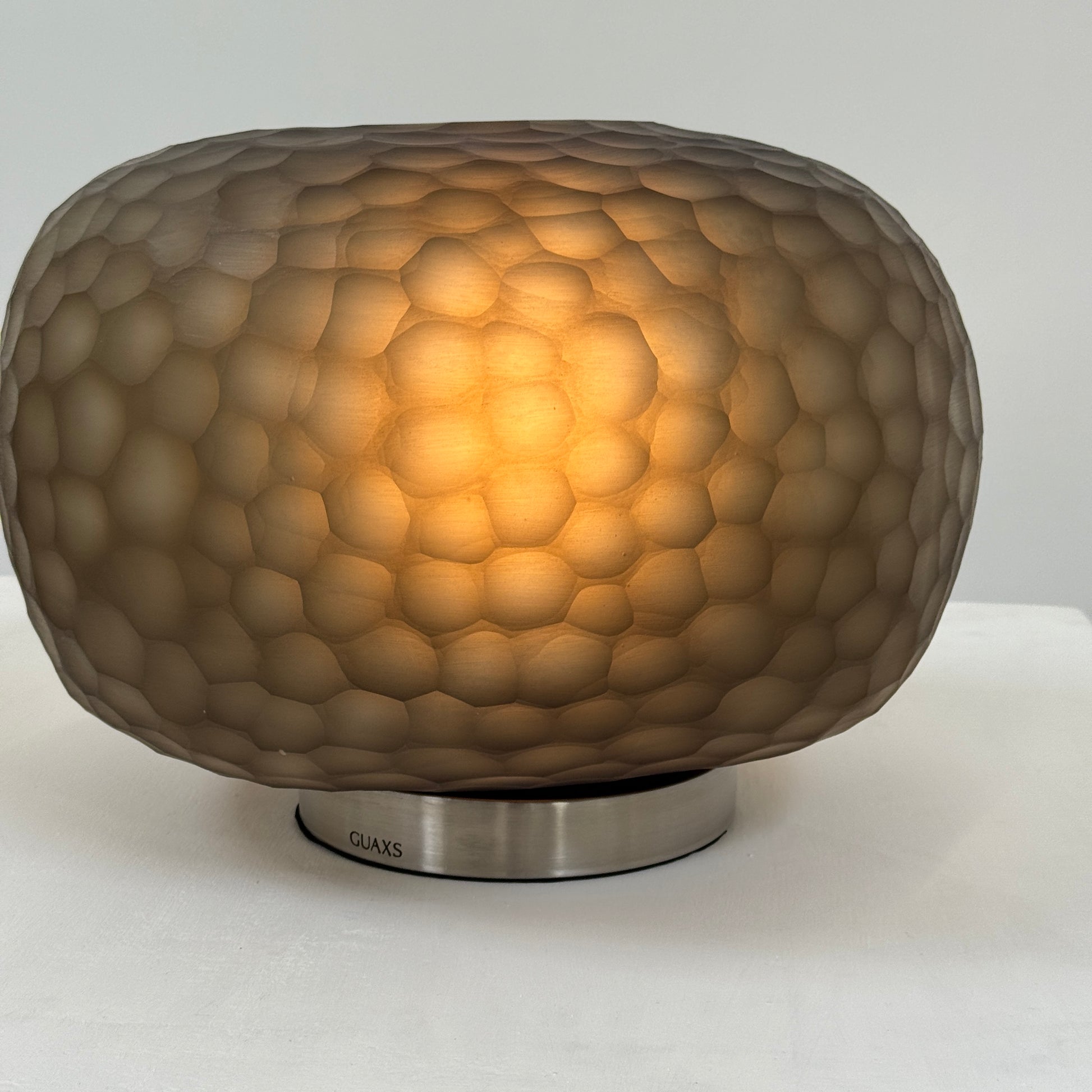 The Guax Erbse 2 Table Lamp by Guax features a mouth-blown, honeycomb-patterned glass shade on a round metal base, with an exposed black power cord for a touch of modern elegance.