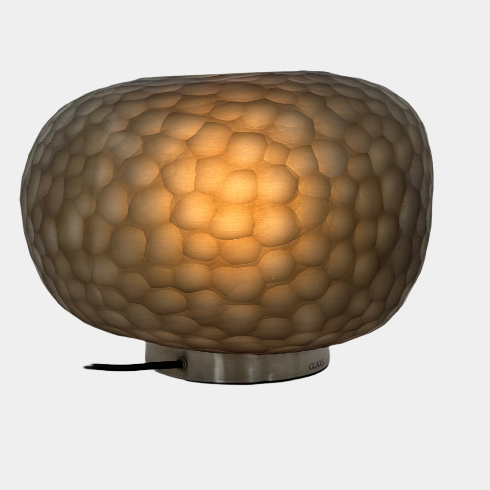 The Guax Erbse 2 Table Lamp by Guax features a mouth-blown, honeycomb-patterned glass shade on a round metal base, with an exposed black power cord for a touch of modern elegance.