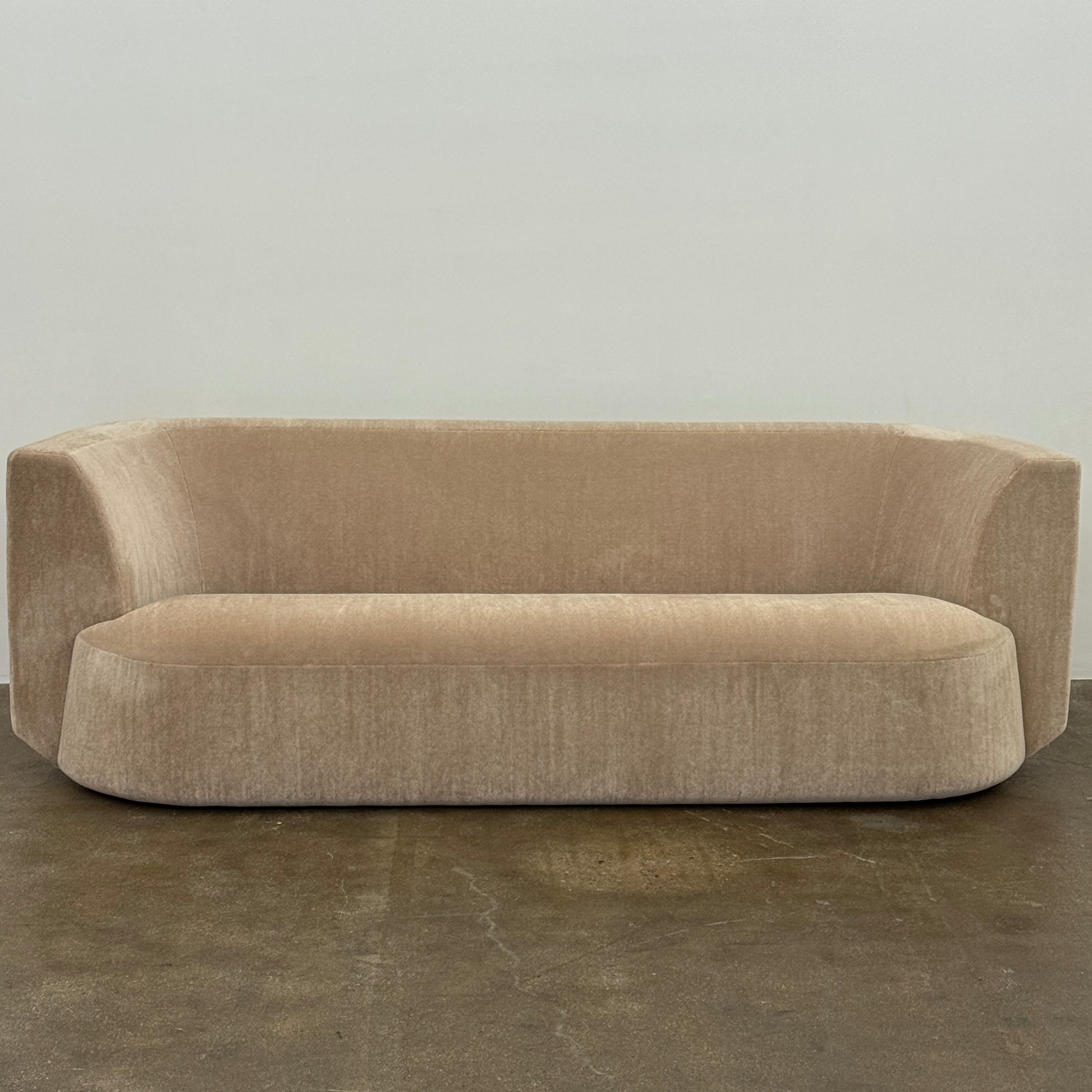 The tan, modern Holly Hunt Caste Malta Sofa with rounded edges sits stylishly on a polished concrete floor against a white wall, showcasing minimalist design inspired by Holly Hunt’s signature aesthetic.