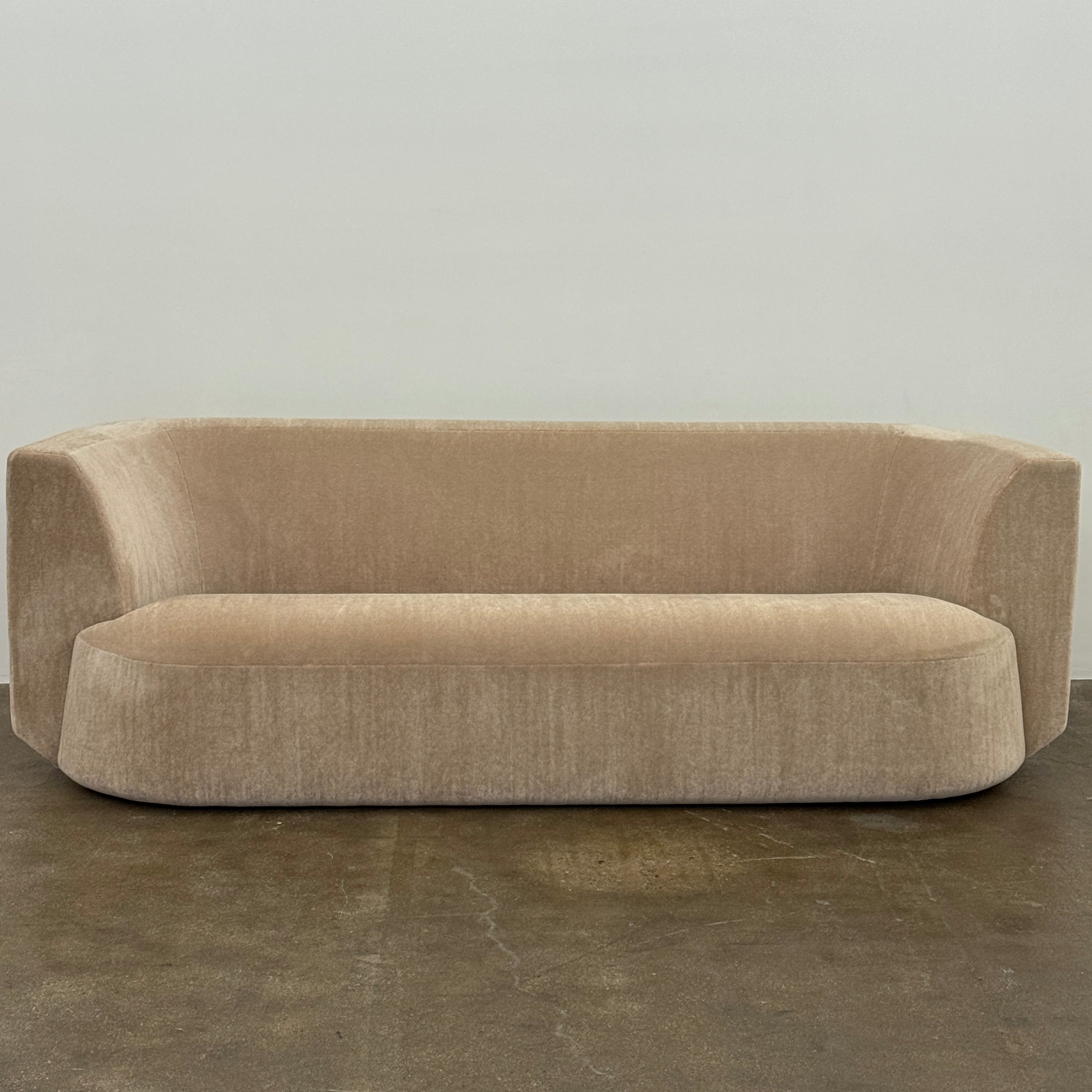 The tan, modern Holly Hunt Caste Malta Sofa with rounded edges sits stylishly on a polished concrete floor against a white wall, showcasing minimalist design inspired by Holly Hunt’s signature aesthetic.